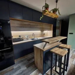 Nowoczesna kuchnia z czarnymi szafkami, drewnianymi blatami i wyspą z hokerami na tle podłogi imitującej drewno. Nad wyspą oryginalne oświetlenie z drewnianego bala z roślinami.