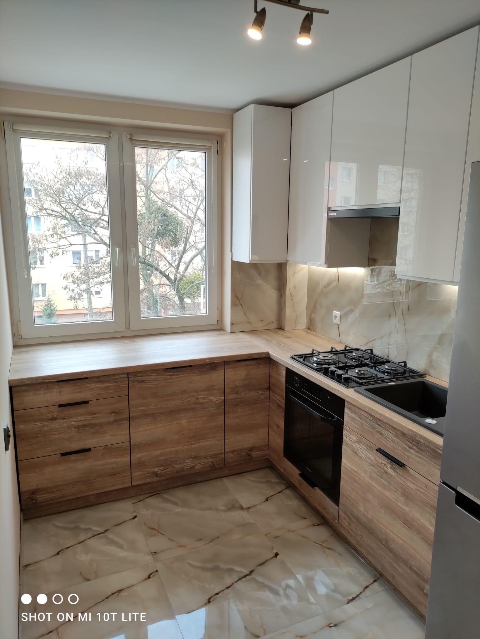 Nowoczesna kuchnia z białymi szafkami górnymi i dolnymi szafkami z drewnopodobnym fornirem, czarnym zlewem i płytą grzewczą, marmurowymi płytkami na podłodze i ścianie nad blatem, widok na drzewo...