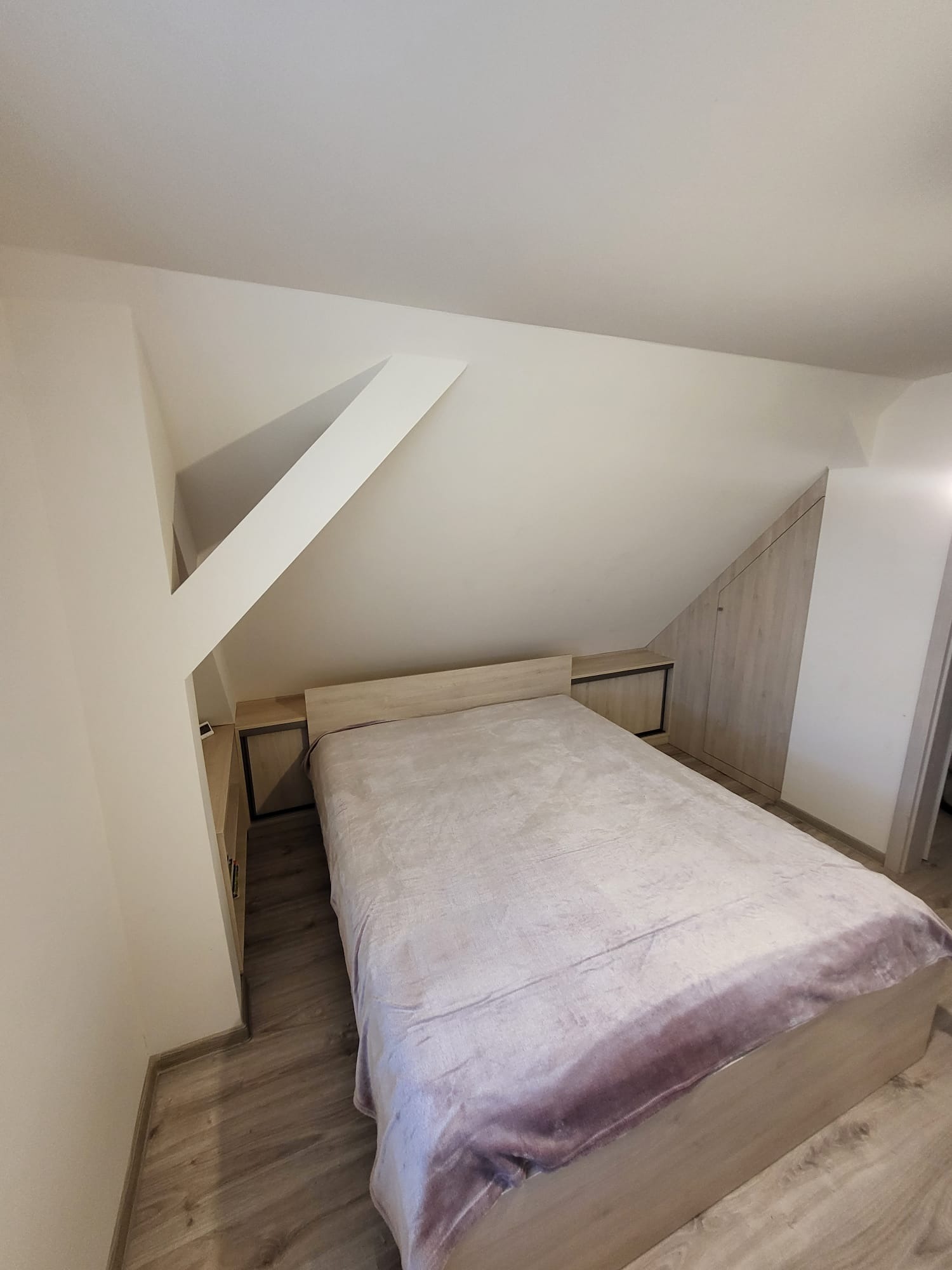 Sypialnia na poddaszu ze skośnym sufitem, łóżko z beżową ramą i fioletowym pledem, szafa wnękowa z jasnego drewna dopasowana do skosu dachu.