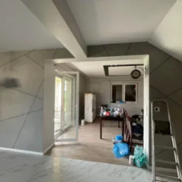 Szara ściana z geometrycznym wzorem w odcieniach szarości, widok na sąsiednie pomieszczenie z zegarem na ścianie, drabina oparta o ścianę, podłoga wyłożona płytkami imitującymi marmur.