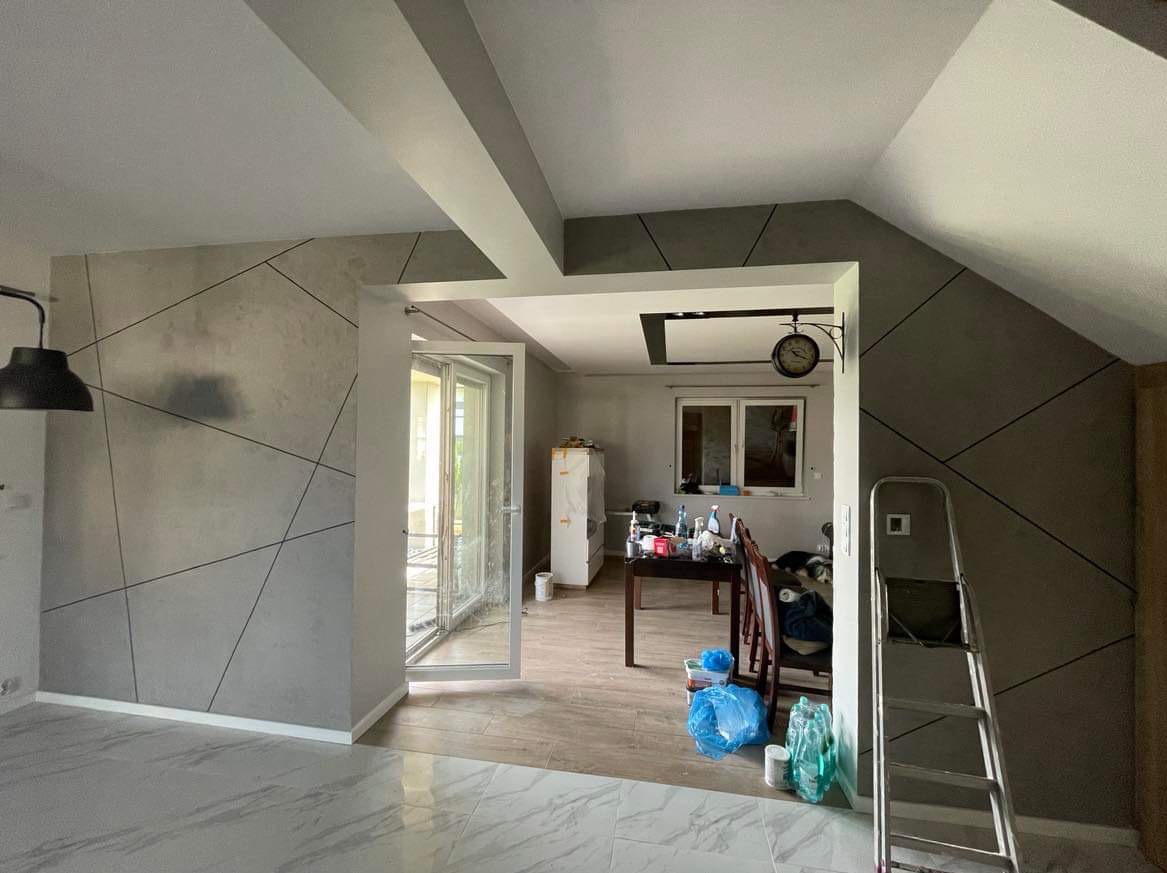 Szara ściana z geometrycznym wzorem w odcieniach szarości, widok na sąsiednie pomieszczenie z zegarem na ścianie, drabina oparta o ścianę, podłoga wyłożona płytkami imitującymi marmur.