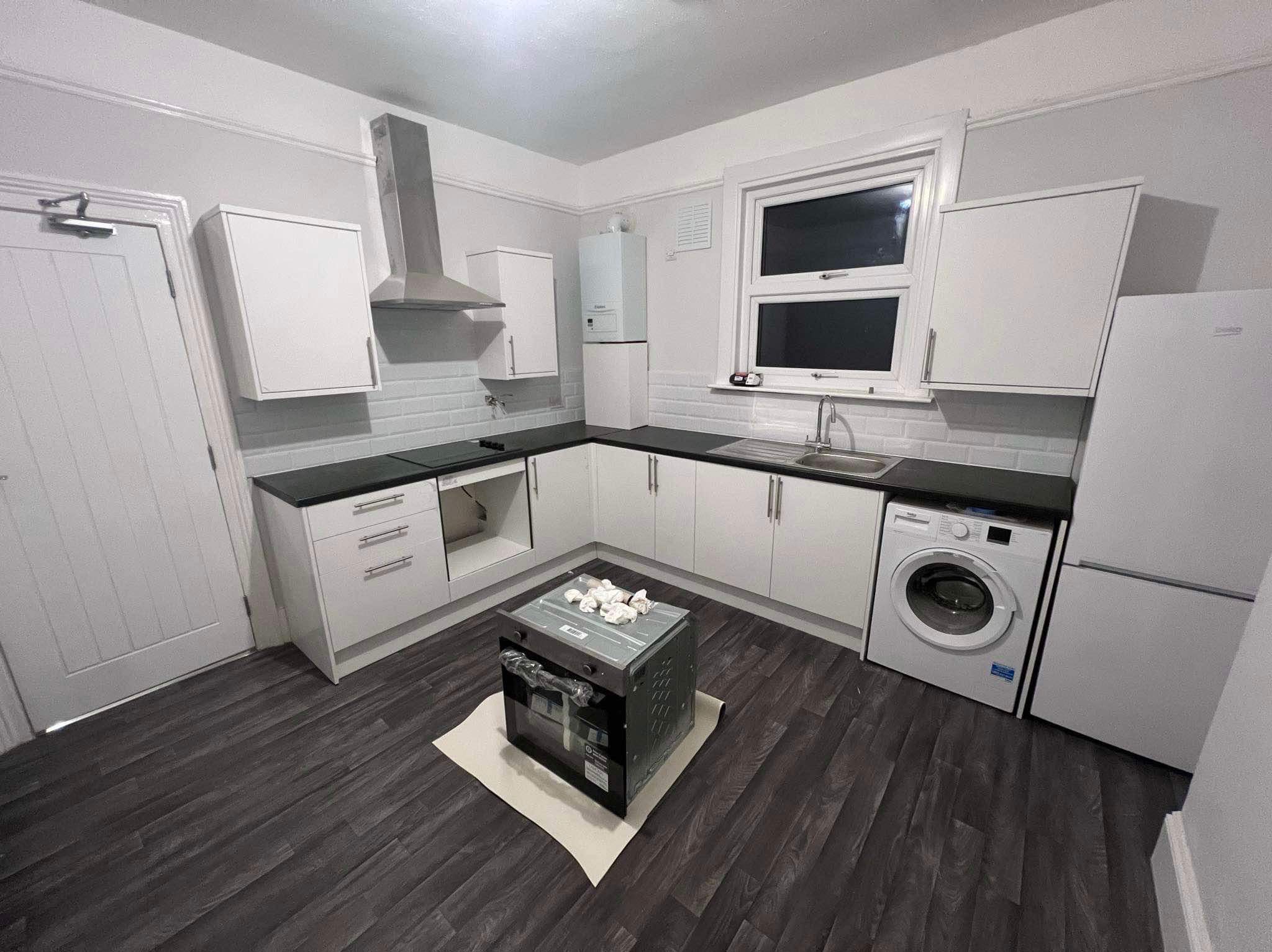 Nowa, jasna kuchnia z białymi szafkami, czarnym blatem i ciemną podłogą; piekarnik na podkładce czeka na montaż na środku pomieszczenia.