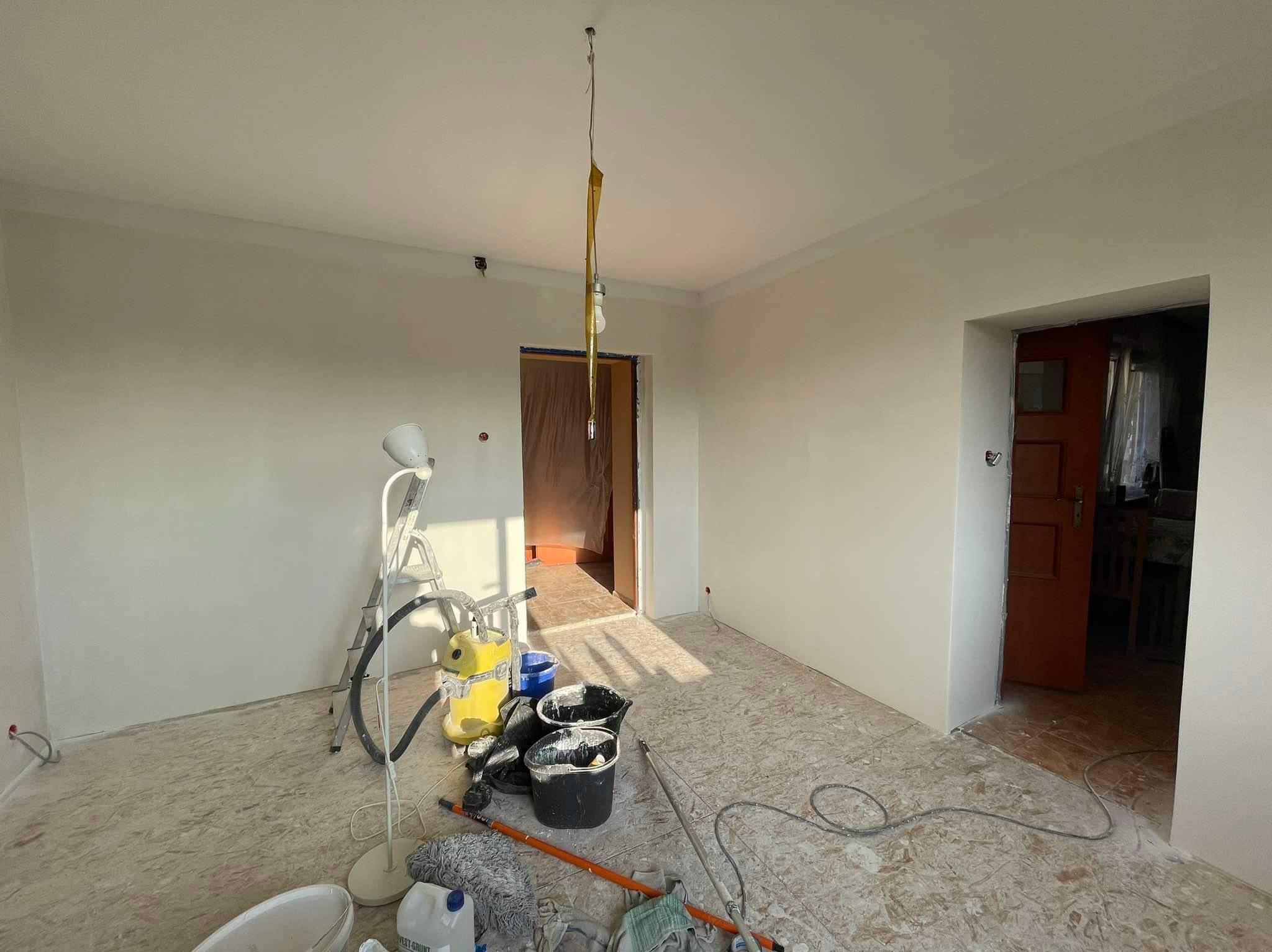 Pokój po malowaniu ścian na jasny odcień, widoczne narzędzia malarskie, drabina i niedoskonałości na podłodze.