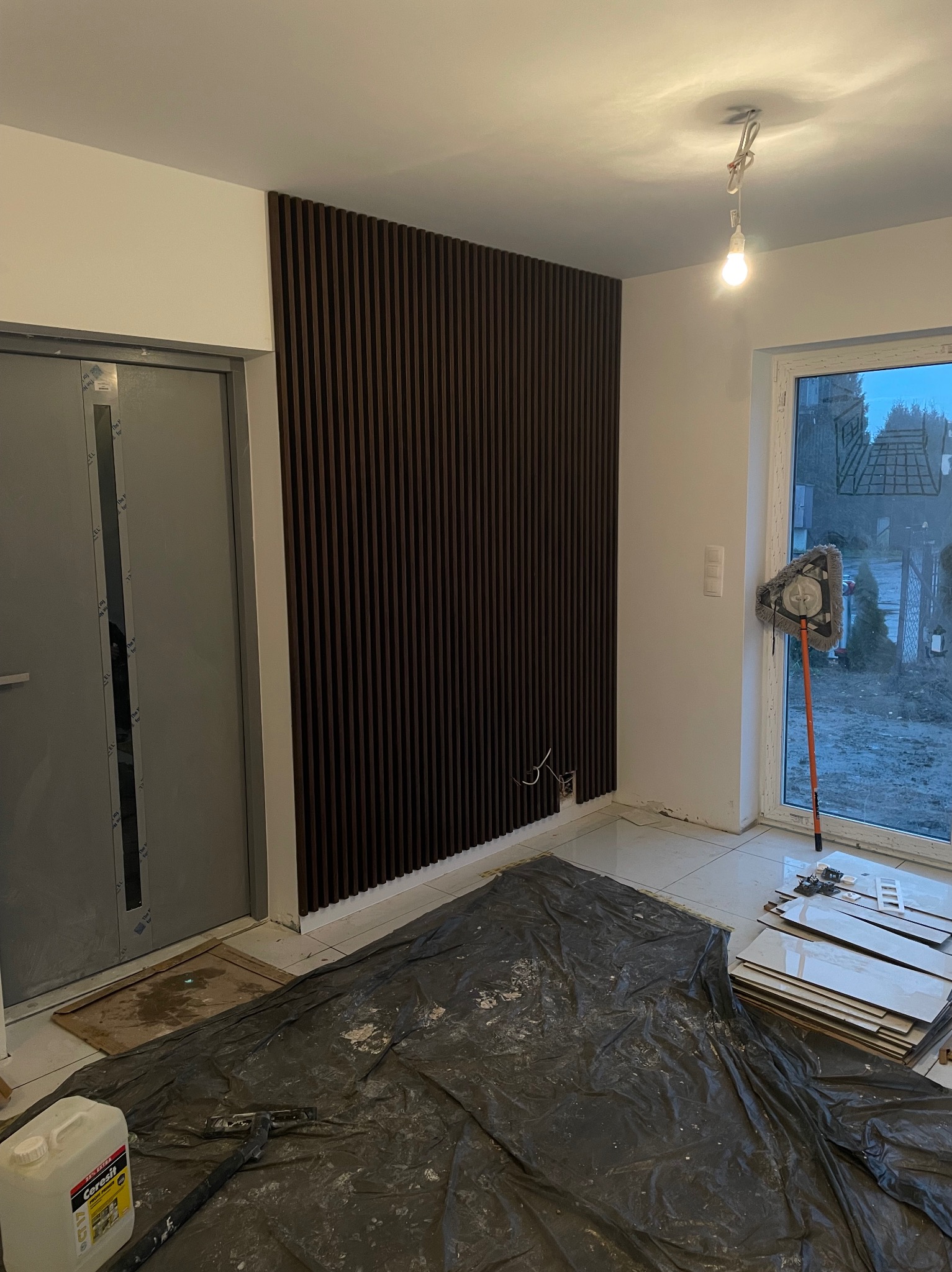 Montaż paneli ściennych z ciemnego drewna na białej ścianie, widoczne przewody elektryczne i przygotowane płytki podłogowe, podłoga zabezpieczona folią ochronną.