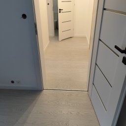 OLEKSANDR SOSKOV - Wnętrze mieszkania z widocznymi trzema białymi drzwiami z czarnymi klamkami, podłoga z jasnych paneli imitujących drewno, widoczne gniazdko elektryczne i otwór na kabel w ścianie.