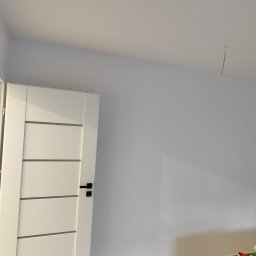 OLEKSANDR SOSKOV - Białe, nowoczesne drzwi wewnętrzne z poziomymi frezowaniami i czarną klamką w nowo wykończonym pokoju w Świnoujściu, widoczny fragment ściany i sufitu z wystającym kablem.