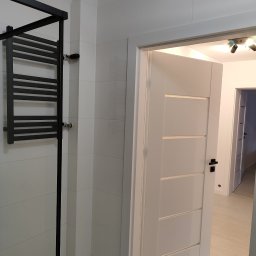 OLEKSANDR SOSKOV - Nowoczesna łazienka z białymi płytkami, czarnym grzejnikiem drabinkowym i otwartymi białymi drzwiami z czarną klamką, widok na jasne wnętrze.