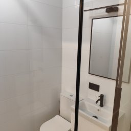 OLEKSANDR SOSKOV - Nowoczesna, minimalistyczna łazienka z białymi płytkami, wiszącą toaletą i szafką z umywalką, czarną armaturą i lustrem w ciemnej ramie, oddzielona szklaną kabiną.