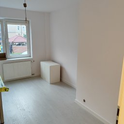 OLEKSANDR SOSKOV - Pomalowane na biało puste pomieszczenie z szarą podłogą imitującą drewno, białym grzejnikiem pod oknem i białą szafką z drzwiczkami w rogu, widoczne ślady po gniazdku elektrycznym na ścianie.