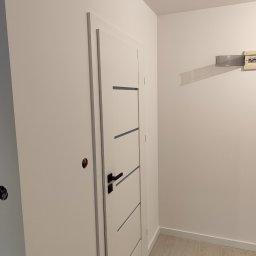 OLEKSANDR SOSKOV - Nowo zamontowane białe drzwi wewnętrzne z czarną klamką i poziomymi frezami, widoczna instalacja elektryczna w ścianie, świeżo pomalowane ściany i jasna podłoga panelowa.