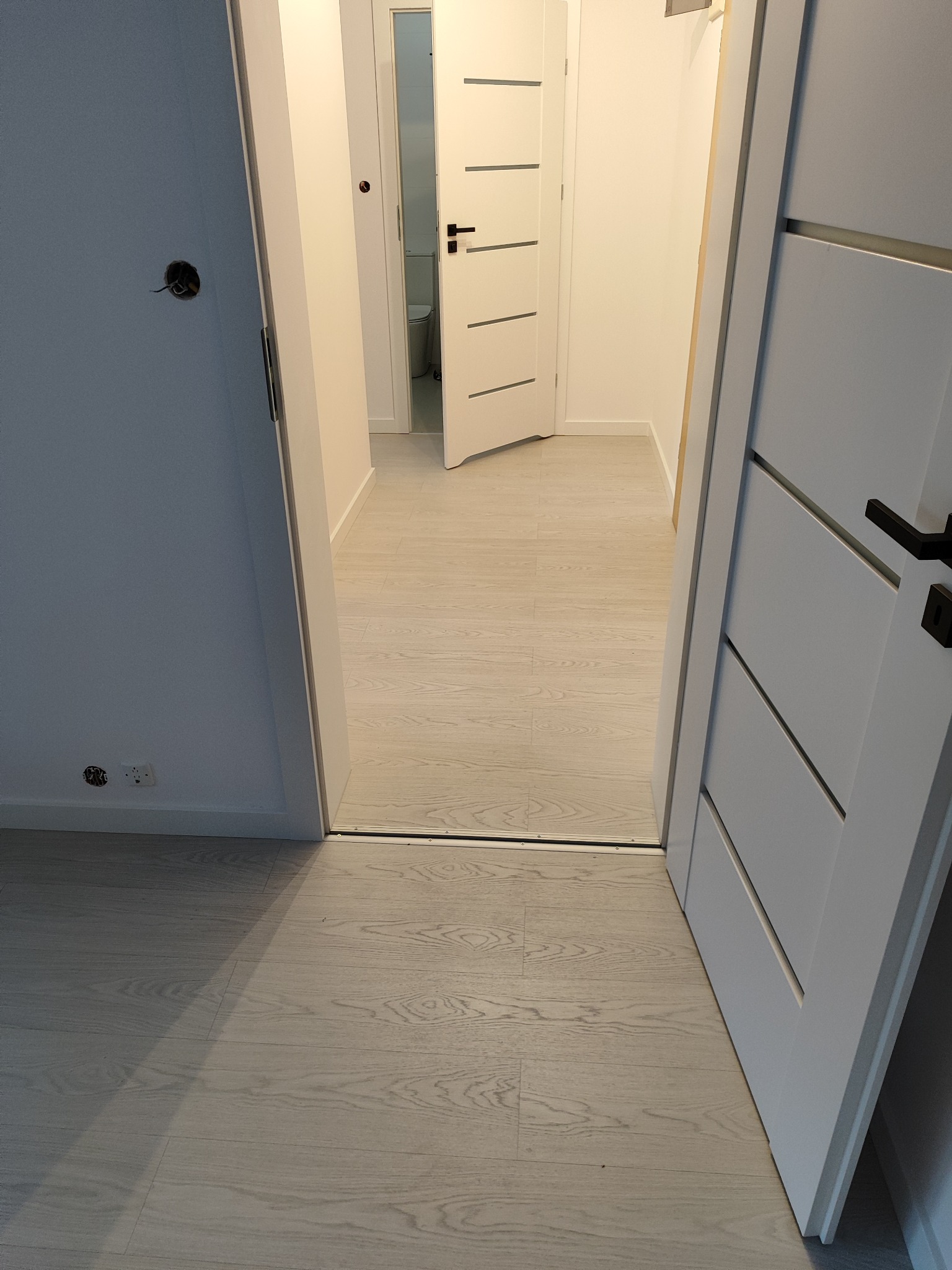 Wnętrze mieszkania z widocznymi trzema białymi drzwiami z czarnymi klamkami, podłoga z jasnych paneli imitujących drewno, widoczne gniazdko elektryczne i otwór na kabel w ścianie.