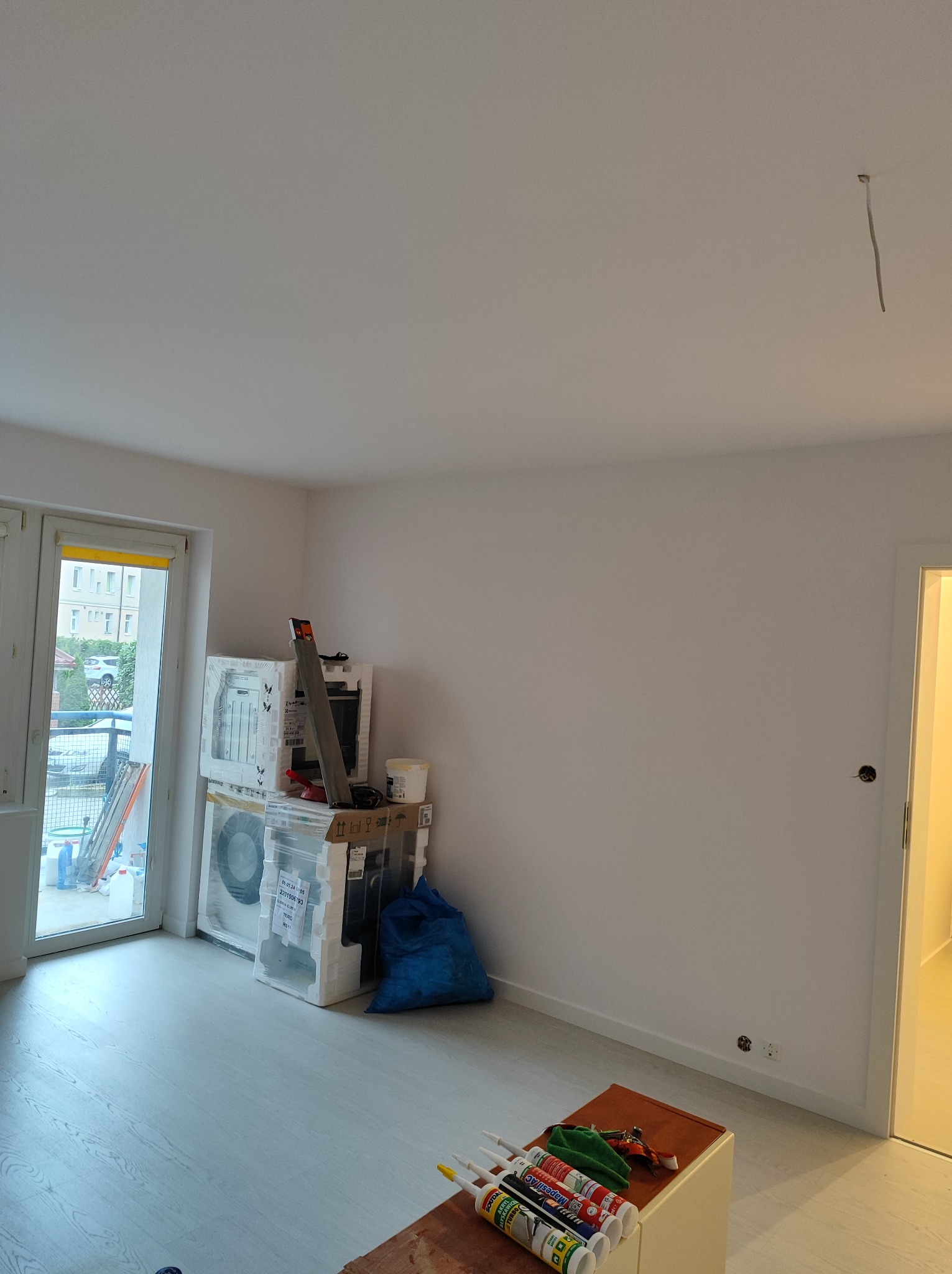 Pokój w trakcie prac przygotowawczych: świeżo pomalowane ściany na biało, zabezpieczone folią sprzęty AGD, widoczne narzędzia i tuby z masami uszczelniającymi na niskim stoliku, gładka, jasna podłoga.