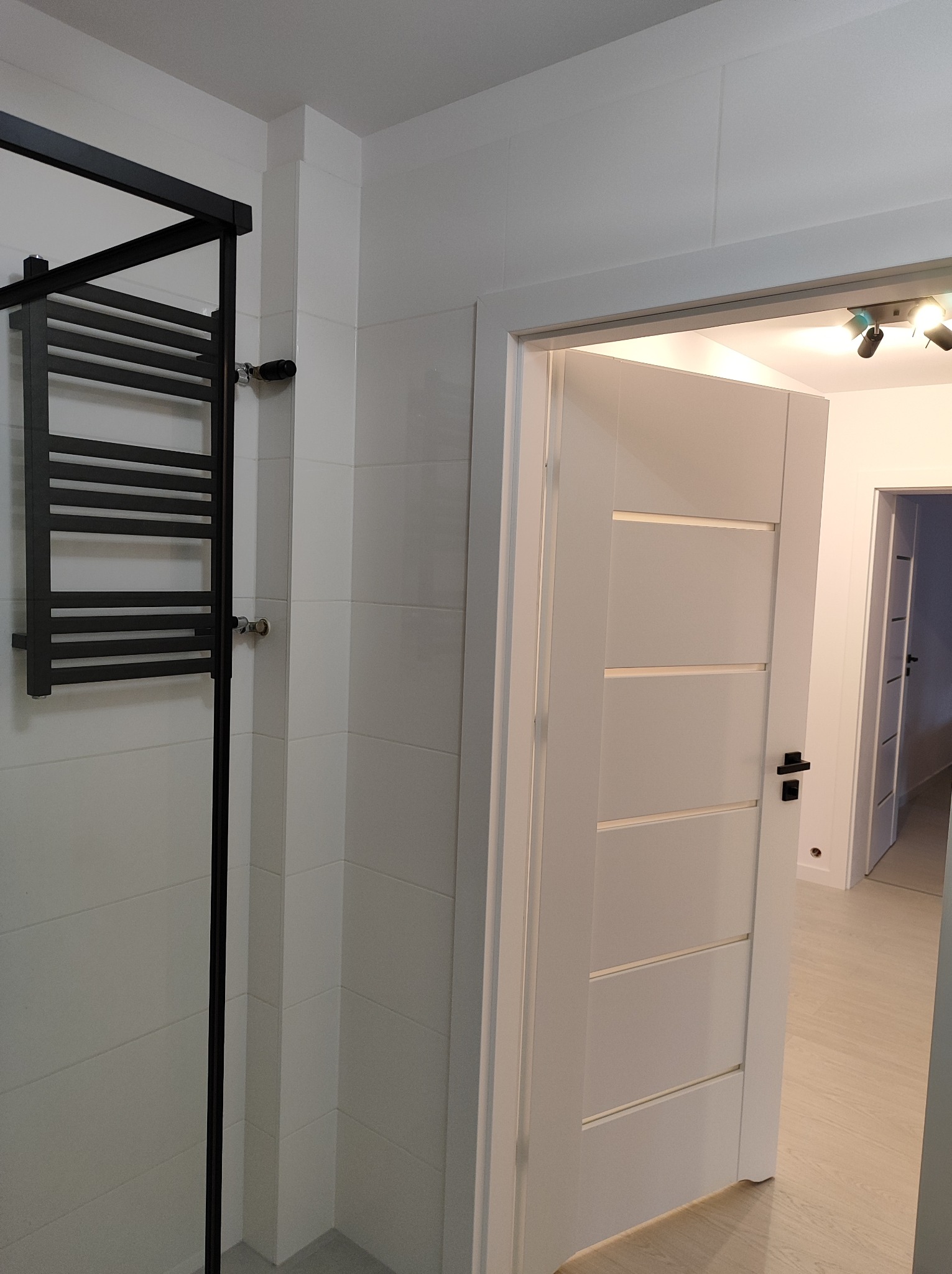 Nowoczesna łazienka z białymi płytkami, czarnym grzejnikiem drabinkowym i otwartymi białymi drzwiami z czarną klamką, widok na jasne wnętrze.