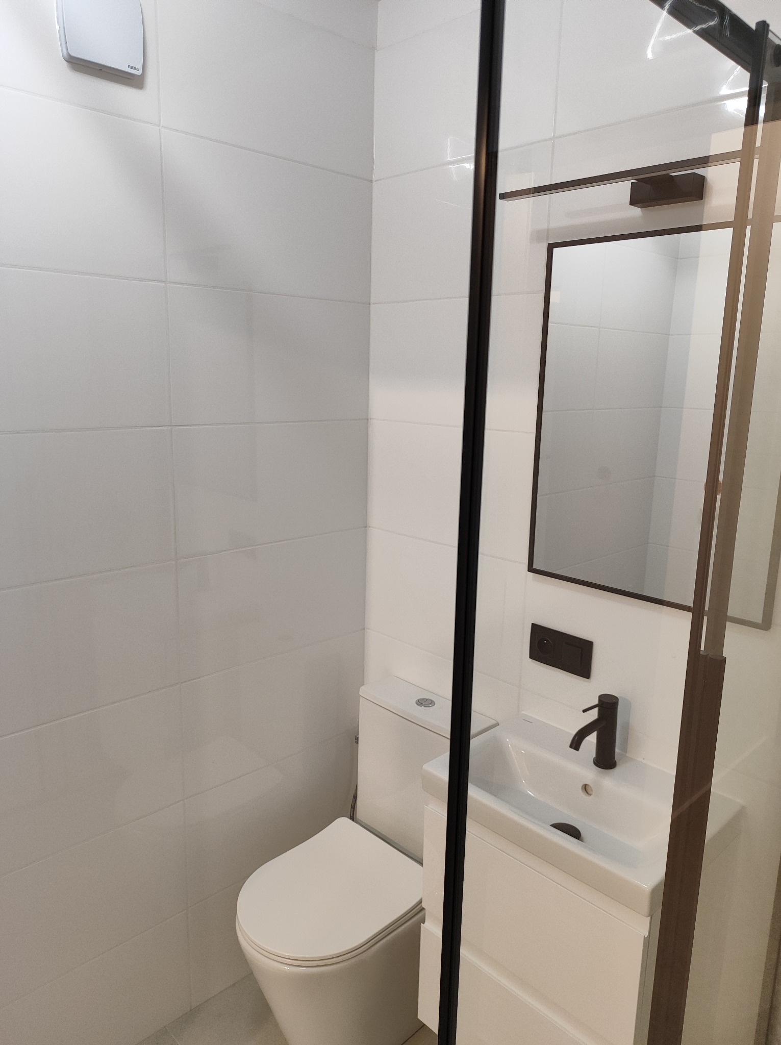 Nowoczesna, minimalistyczna łazienka z białymi płytkami, wiszącą toaletą i szafką z umywalką, czarną armaturą i lustrem w ciemnej ramie, oddzielona szklaną kabiną.