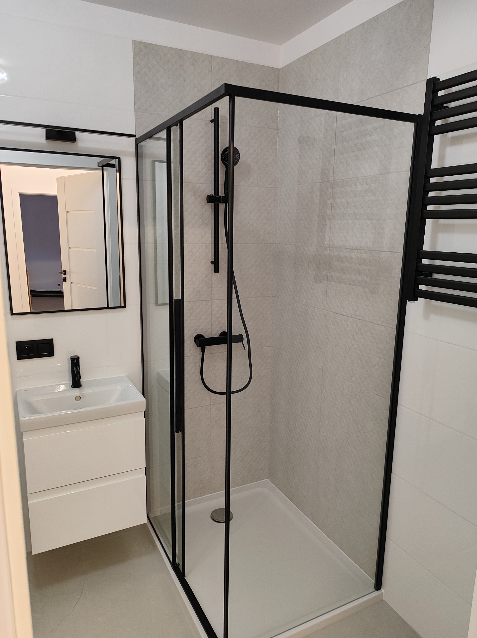 Nowoczesna łazienka z kabiną prysznicową typu walk-in z czarnymi profilami, białą umywalką nablatową i szarymi płytkami imitującymi kamień.