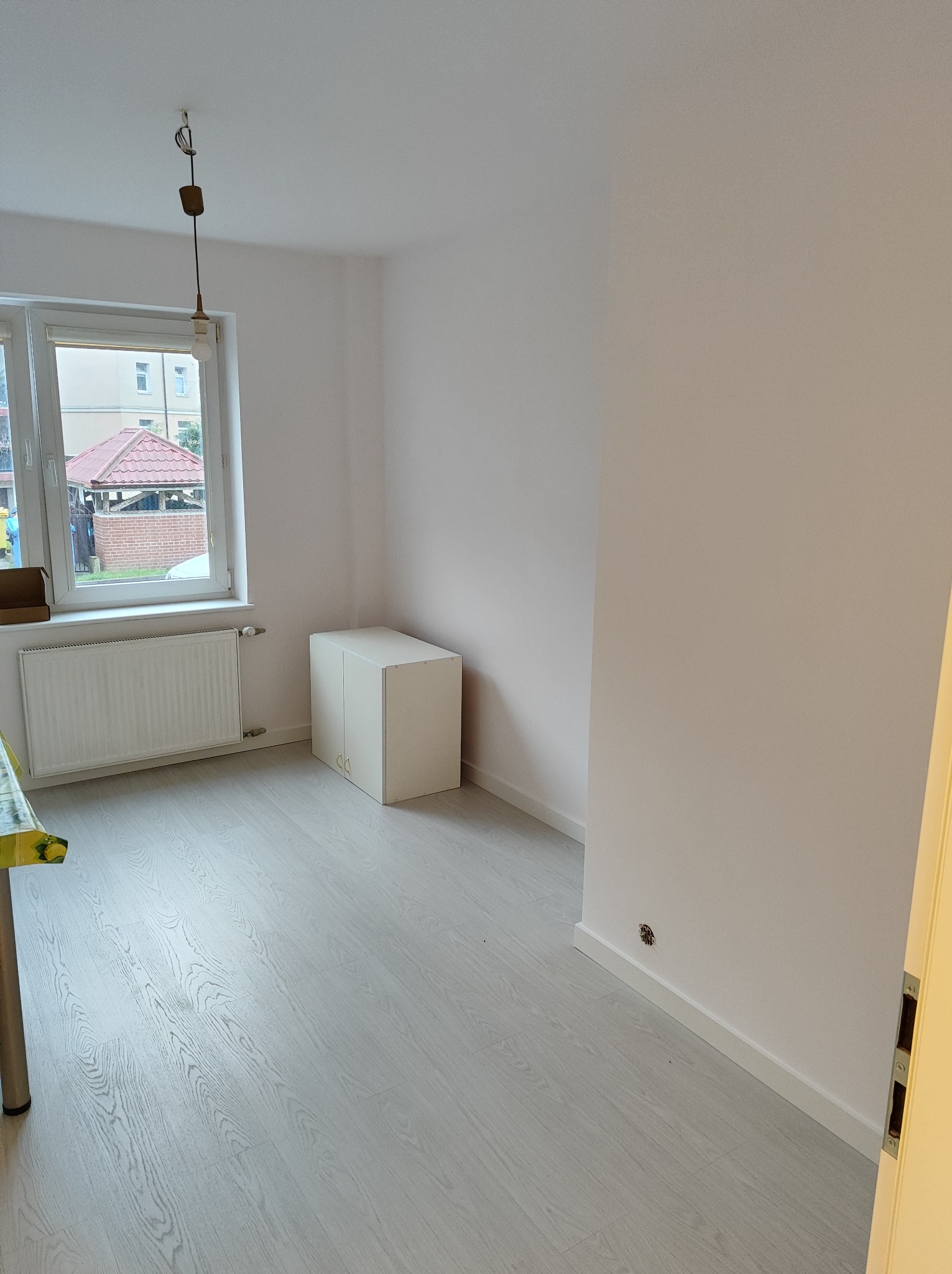 Pomalowane na biało puste pomieszczenie z szarą podłogą imitującą drewno, białym grzejnikiem pod oknem i białą szafką z drzwiczkami w rogu, widoczne ślady po gniazdku elektrycznym na ścianie.