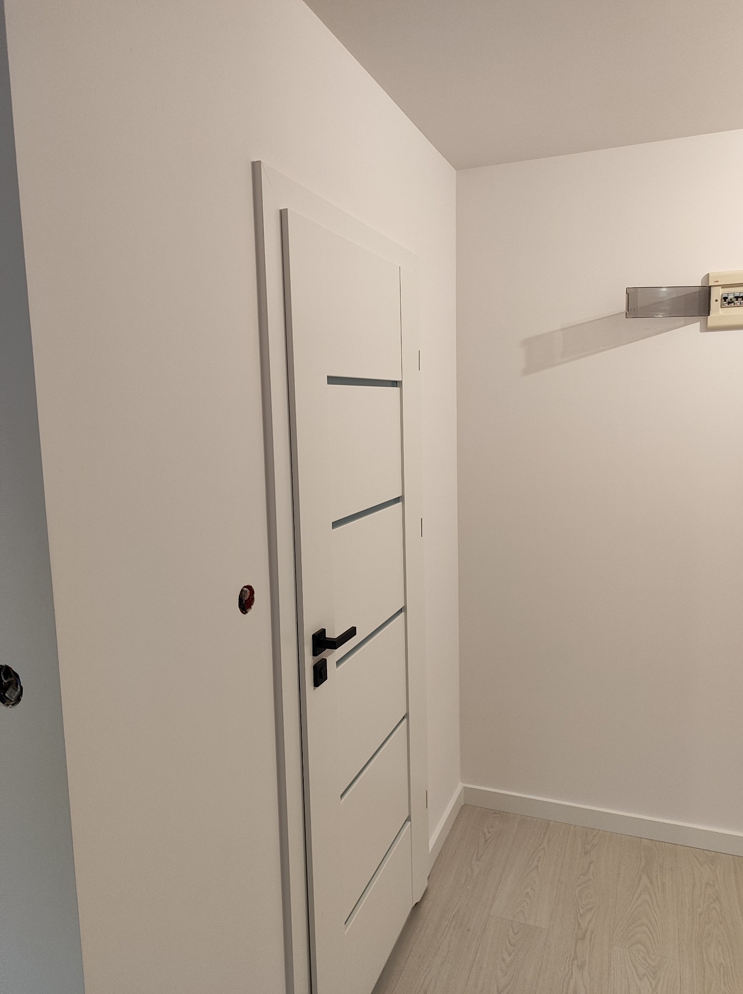 Nowo zamontowane białe drzwi wewnętrzne z czarną klamką i poziomymi frezami, widoczna instalacja elektryczna w ścianie, świeżo pomalowane ściany i jasna podłoga panelowa.