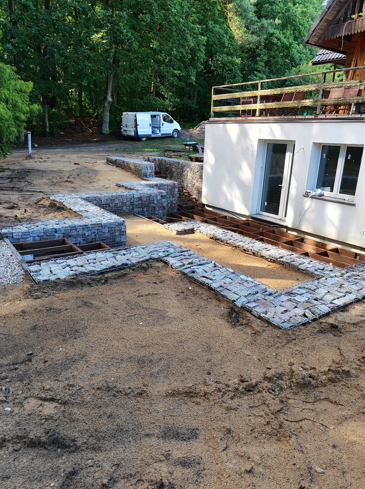 Przygotowanie fundamentów pod taras z gabionów, widoczne drewniane legary i siatka wypełniona kamieniami, biały bus w tle, etap budowy przy domu.