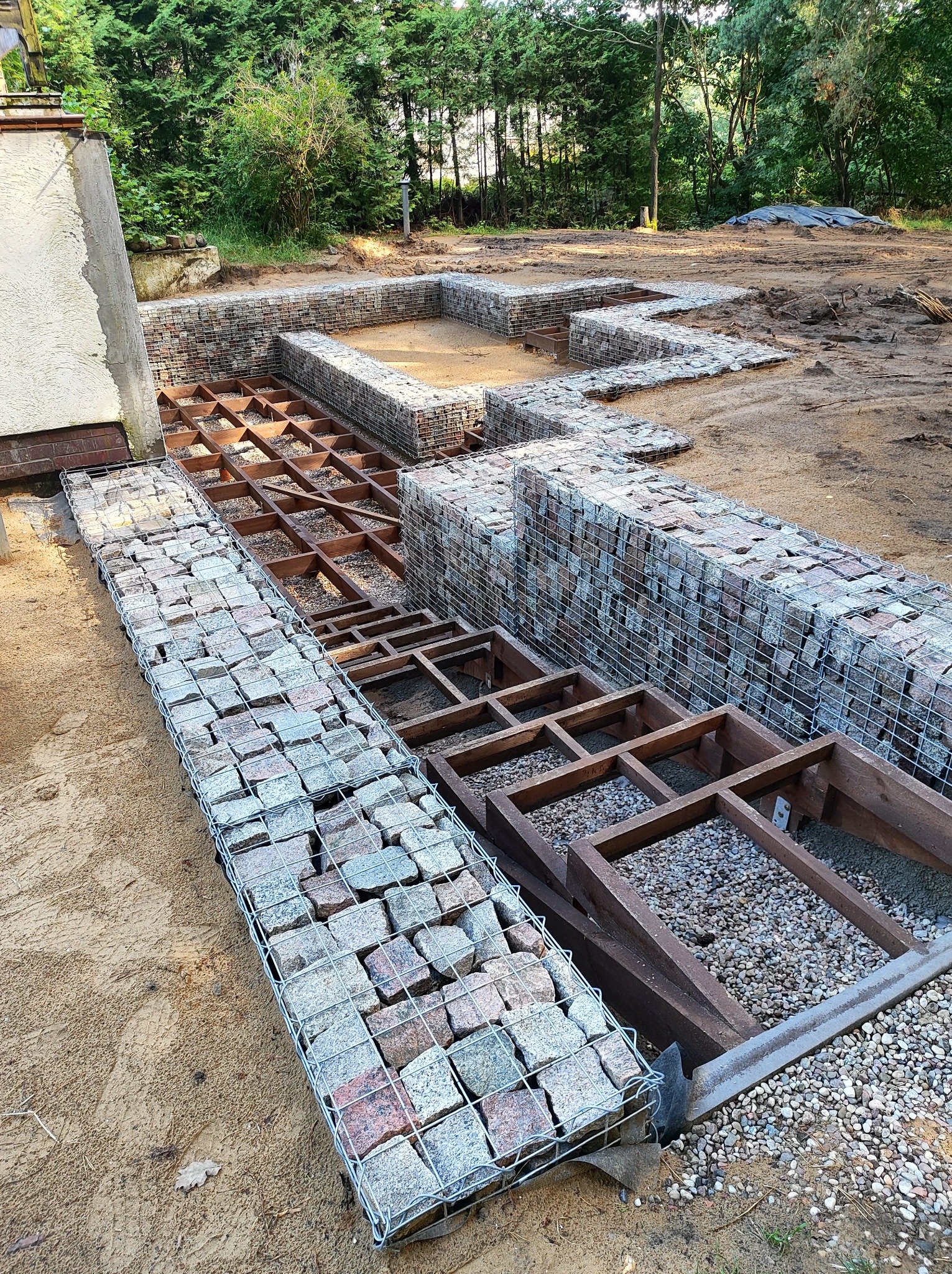 Fundamenty pod taras: drewniana konstrukcja kratowa wypełniona żwirem, otoczona gabionami z kamienia naturalnego, na piaszczystym podłożu.