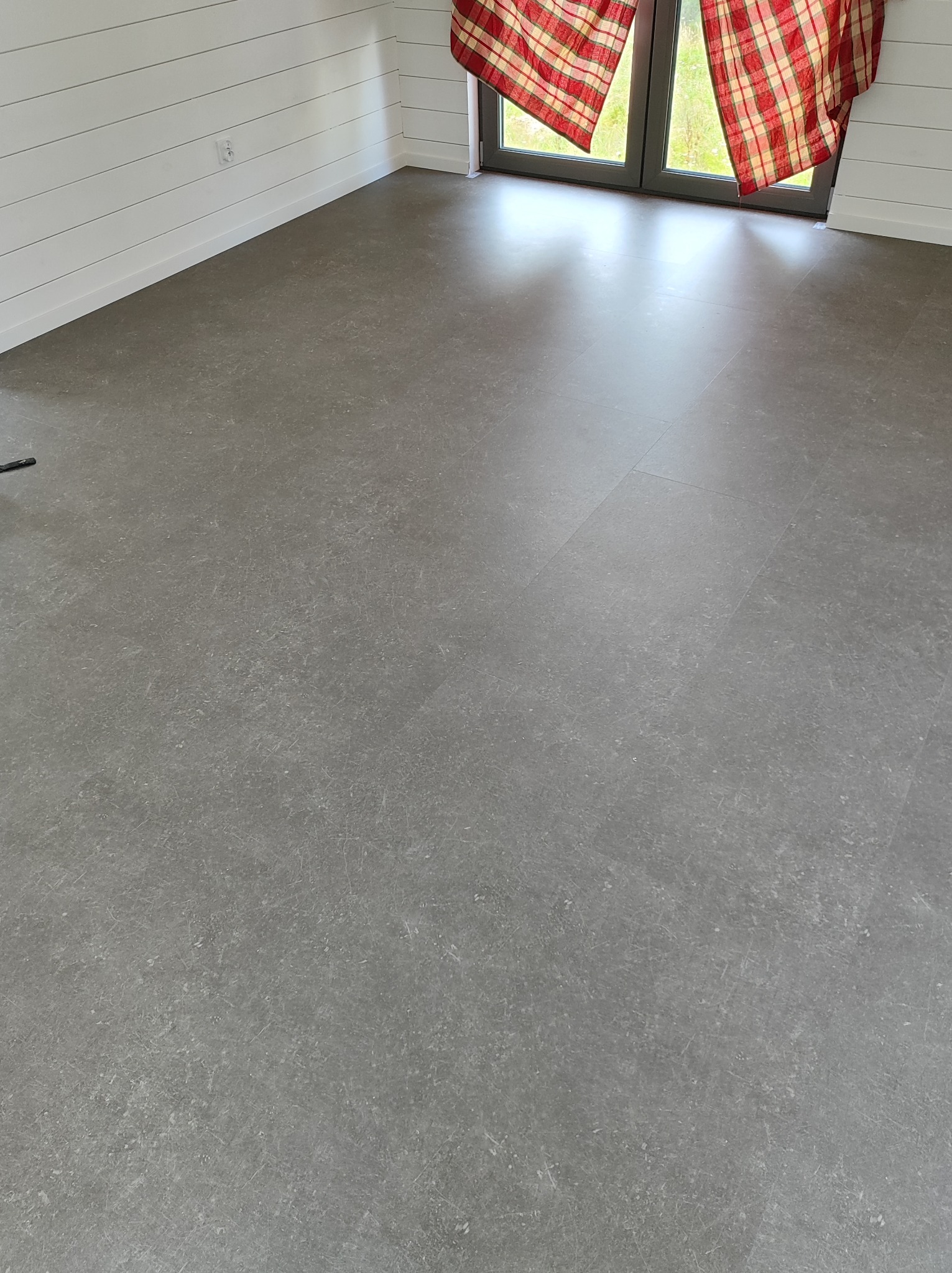 Szara podłoga z paneli winylowych imitujących beton w nowym pomieszczeniu z białymi, poziomymi panelami ściennymi i oknami z czerwoną, kraciastą zasłoną.