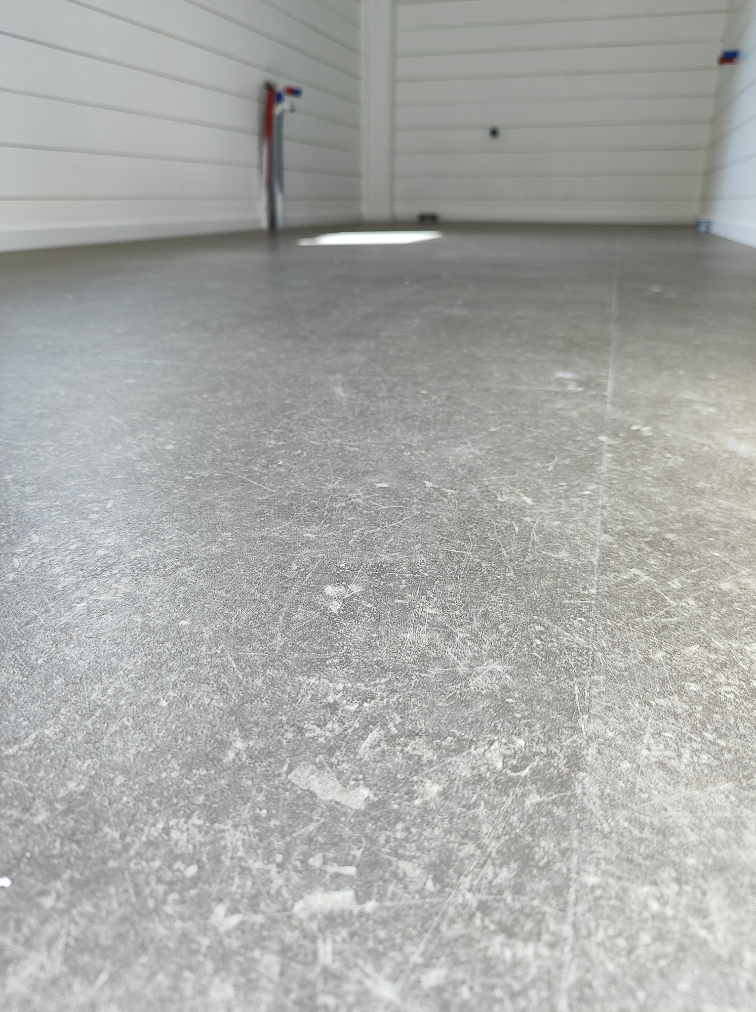 Szara, betonowa wylewka podłogowa w trakcie schnięcia, z widocznymi śladami szlifowania, przy białej ścianie z boazerią i wystającymi rurami instalacji wodnej.