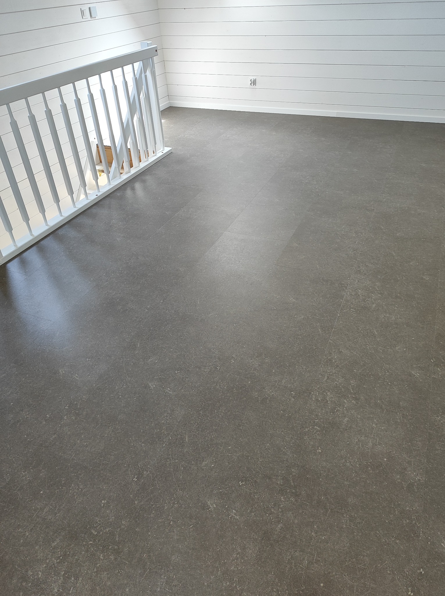 Szara, matowa podłoga z paneli winylowych w nowym pomieszczeniu z białą boazerią i białą balustradą schodów.