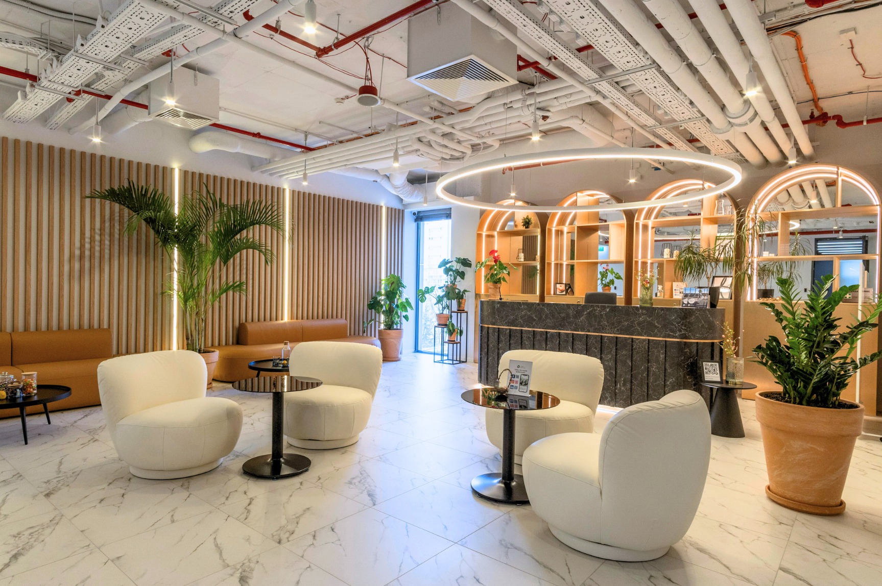 Nowoczesna przestrzeń recepcyjna z marmurową podłogą, białymi fotelami, drewnianymi panelami ściennymi i roślinami doniczkowymi, oświetlona okrągłymi lampami sufitowymi.