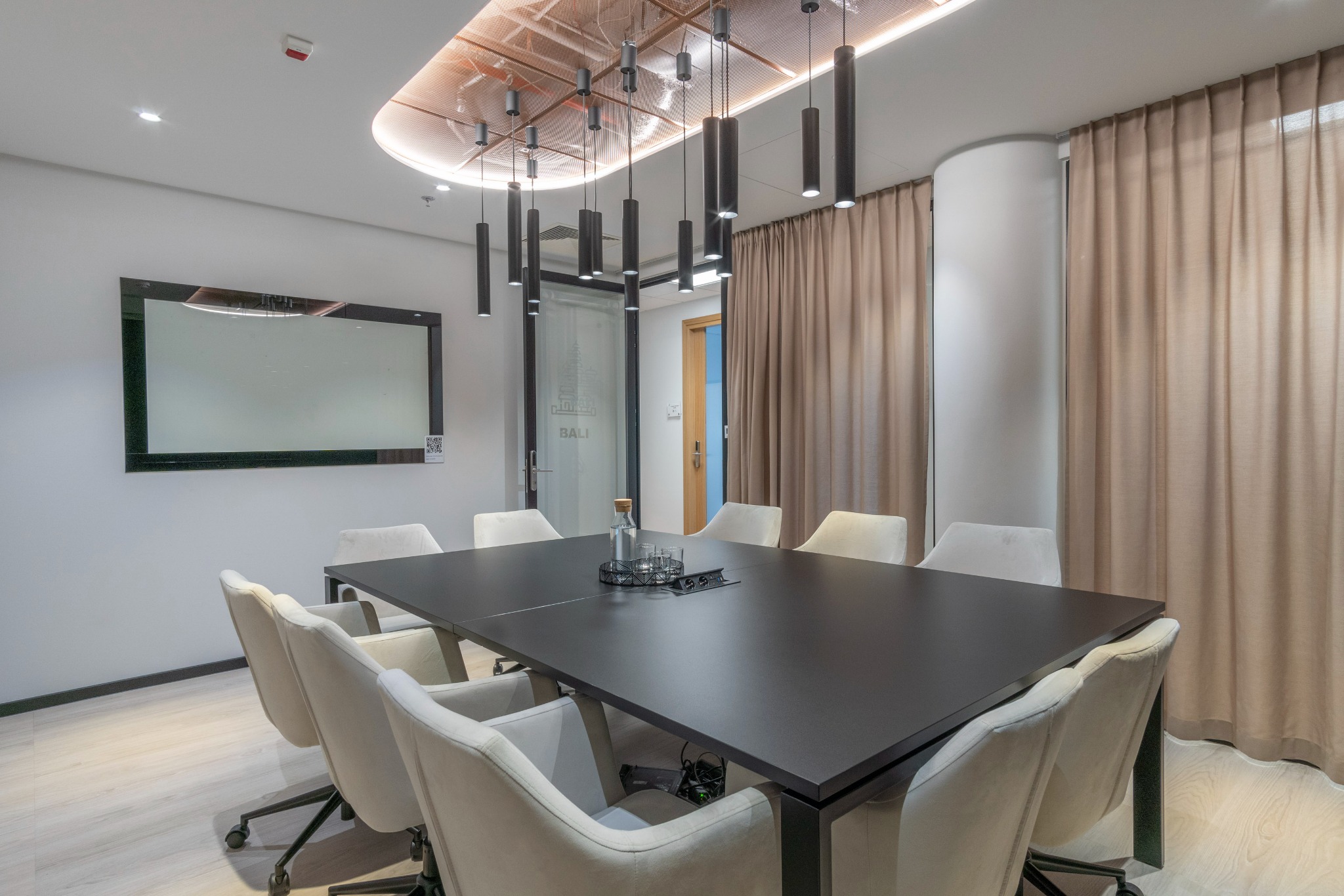 Nowoczesna sala konferencyjna z czarnym stołem, kremowymi krzesłami, szklaną butelką z wodą i wiszącymi lampami cylindrycznymi, widok na szklane drzwi z napisem 'BALI'.