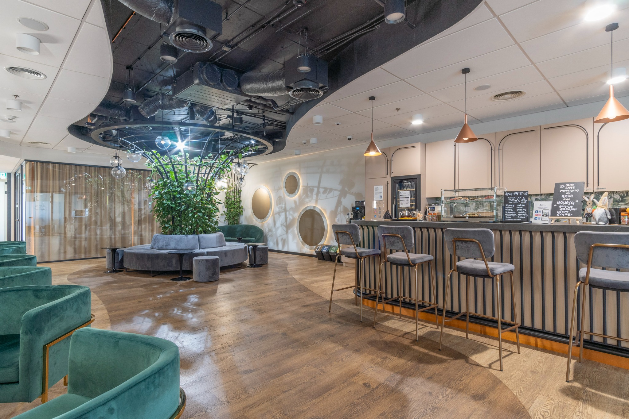 Nowoczesna przestrzeń biurowa z zielonymi akcentami: bar z wysokimi krzesłami, okrągła sofa z roślinami w metalowej konstrukcji, drewniana podłoga i designerskie oświetlenie.