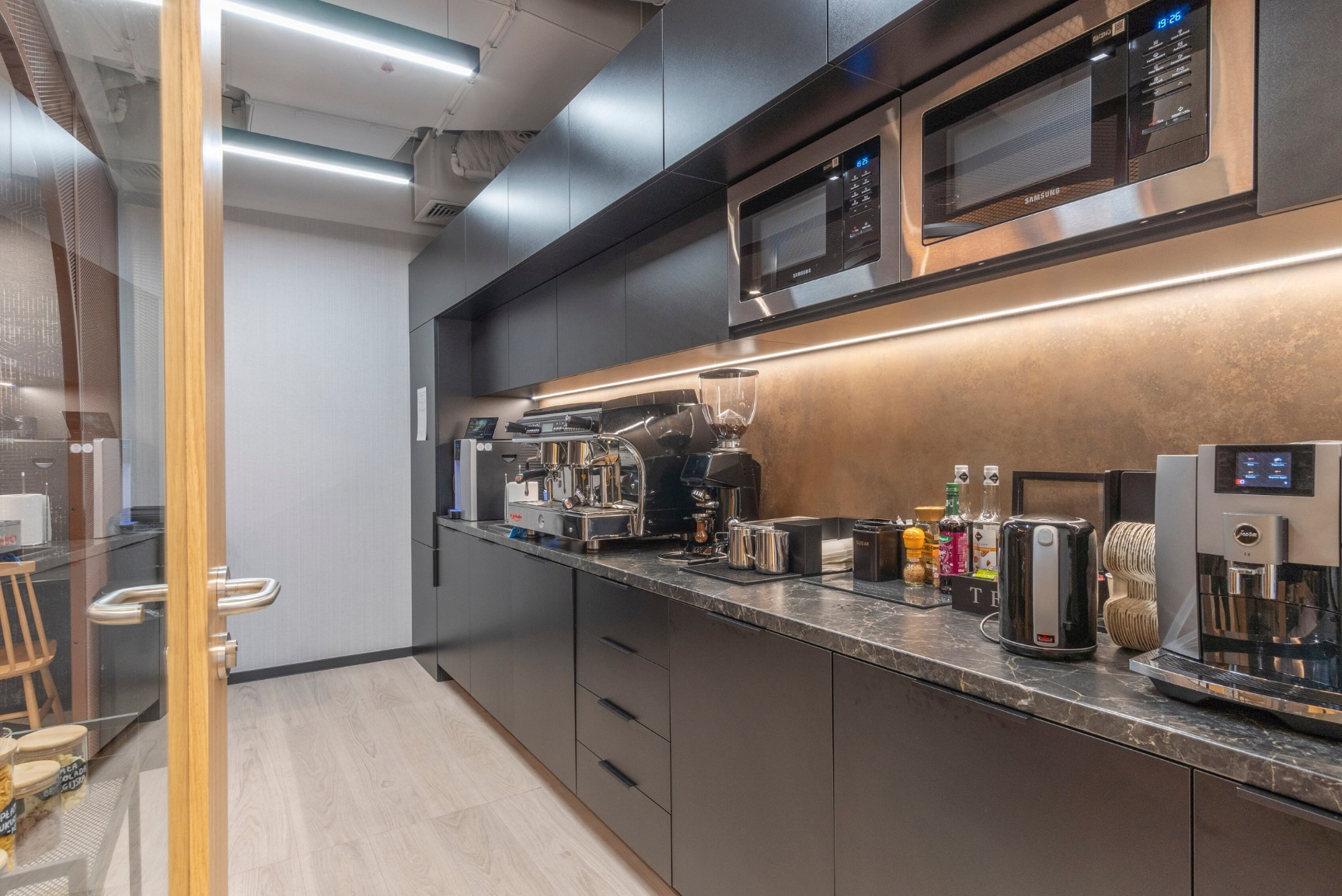 Nowoczesna przestrzeń socjalna w biurze: czarne szafki, marmurowy blat, ekspresy do kawy, mikrofalówki i oświetlenie LED.