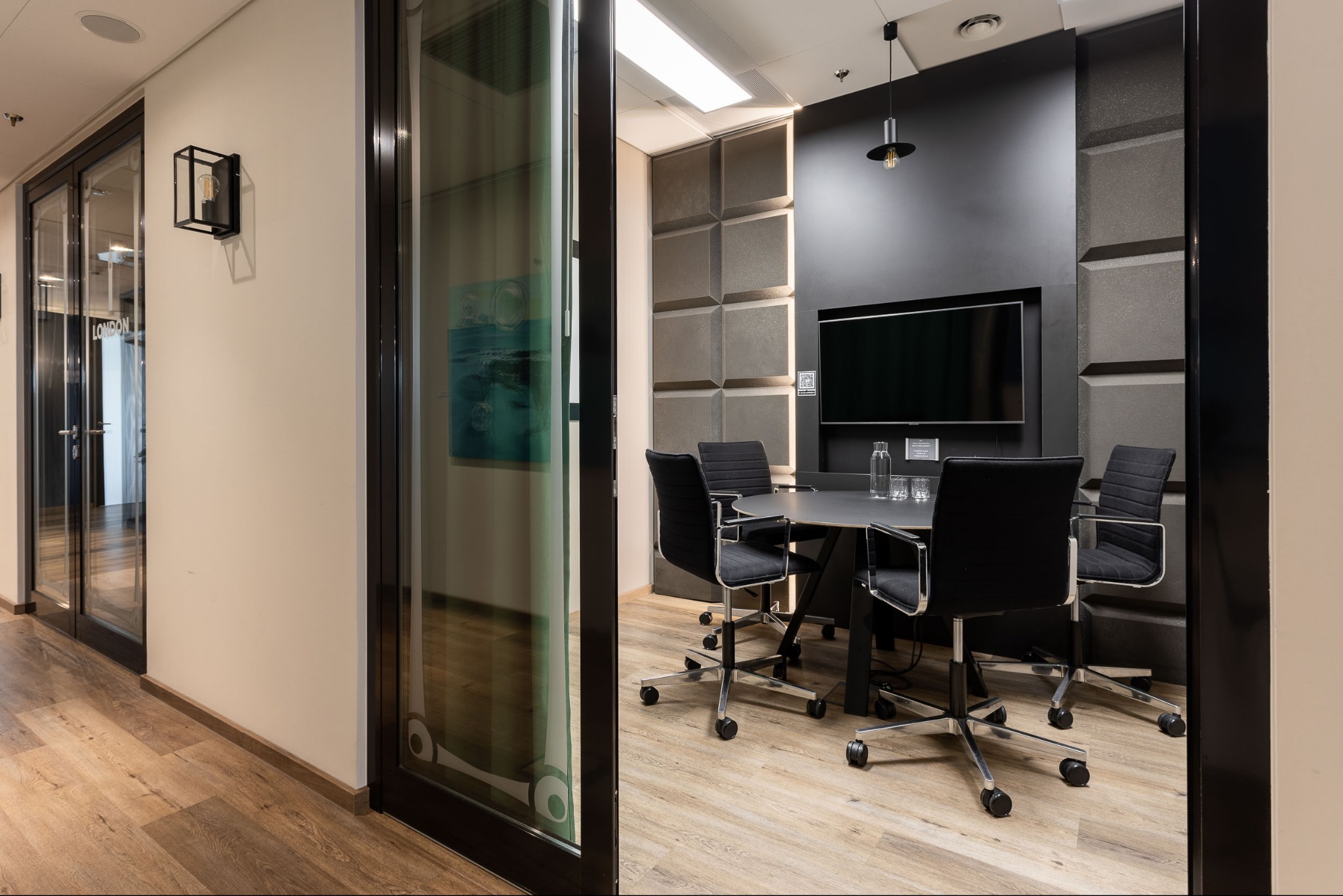 Nowoczesna sala konferencyjna z czarnym stołem i krzesłami na kółkach, podświetlana ściana z paneli akustycznych, przeszklone drzwi z dekoracyjnym motywem.