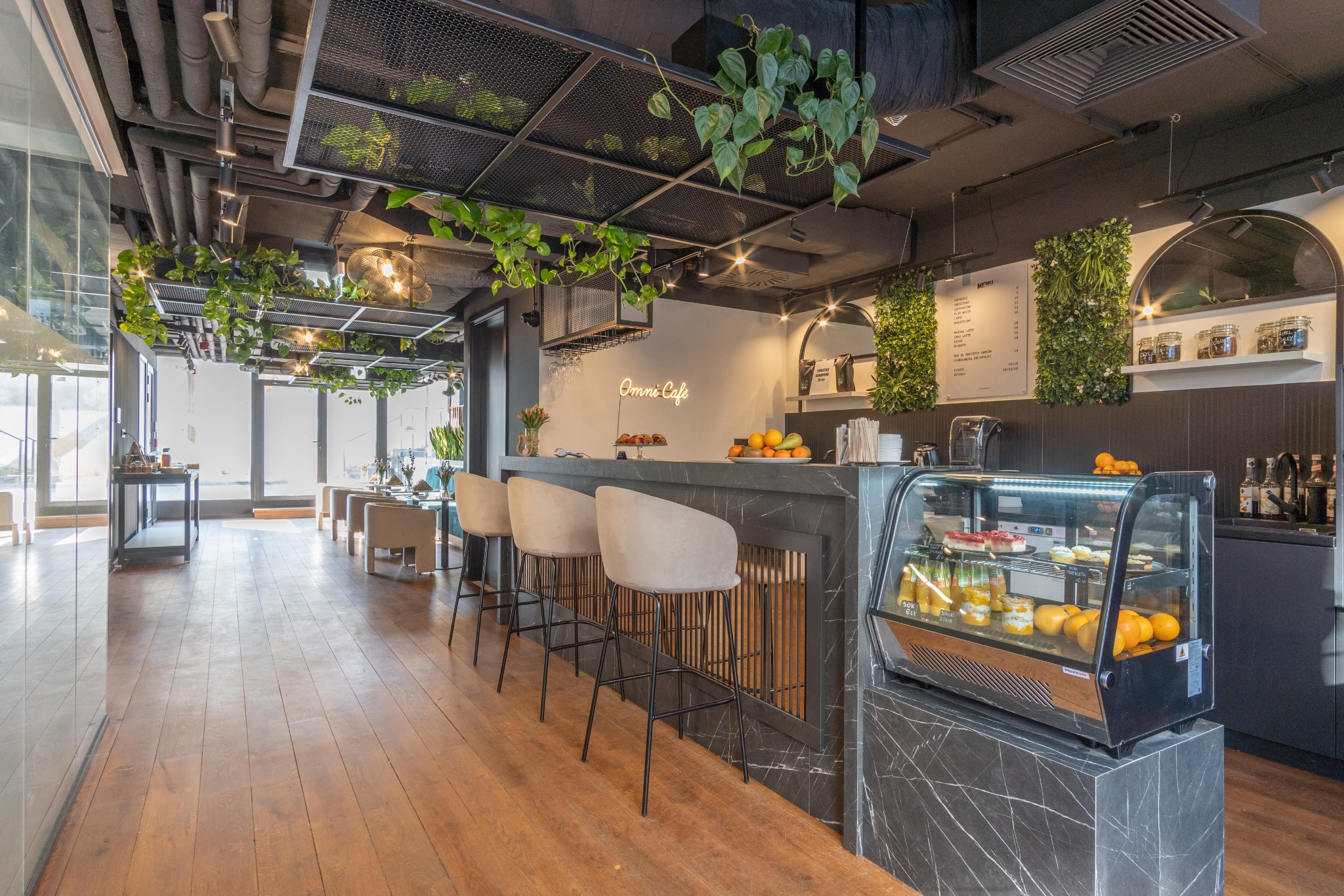 Nowoczesne wnętrze kawiarni z marmurowym barem, hokerami, witryną chłodniczą z deserami i owocami, oraz roślinami zwisającymi z sufitu.