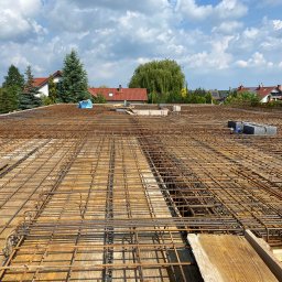 Elbud - Zbrojenie stropu na deskowaniu, widok z góry na przygotowaną konstrukcję z prętów stalowych, w tle zabudowa jednorodzinna i błękitne niebo z chmurami.