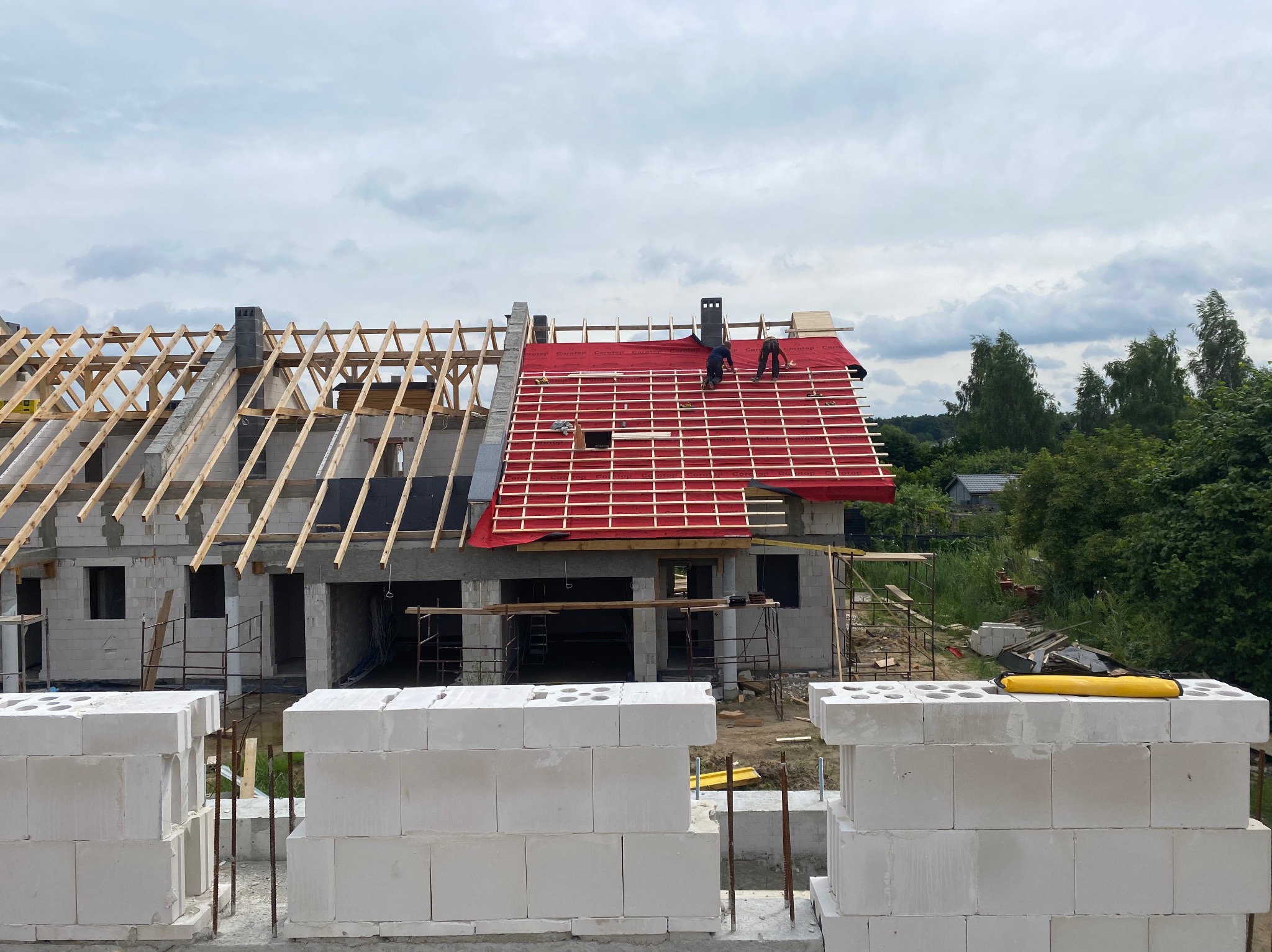 Budowa domu murowanego w Szczecinie, widok na wznoszone ściany z bloczków oraz konstrukcję dachu z czerwoną membraną, na której pracują dwie osoby.