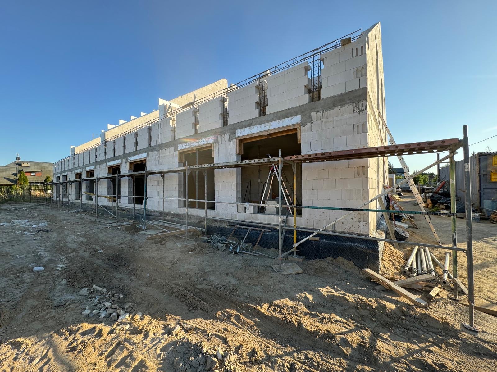 Surowy stan budynku z betonu komórkowego w trakcie budowy, widoczne rusztowania i niezabudowane otwory okienne, teren budowy z piaskiem i gruzem.