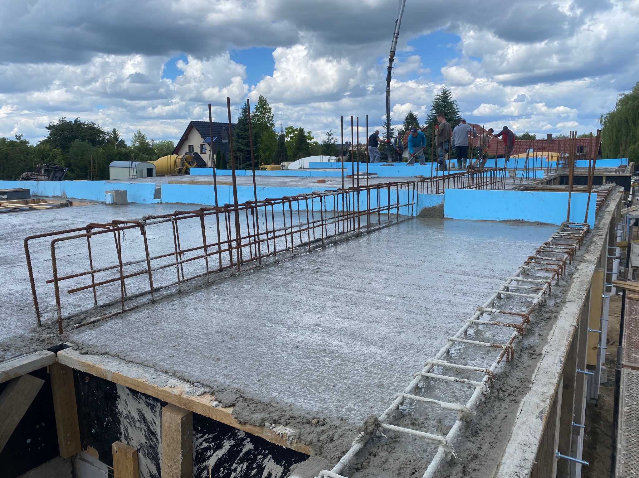 Zalewanie betonem zbrojonej płyty fundamentowej, widoczna szalunek, stalowe pręty zbrojeniowe, izolacja termiczna z niebieskiego styropianu, ekipa budowlana w tle, pompa do betonu i betoniarka.