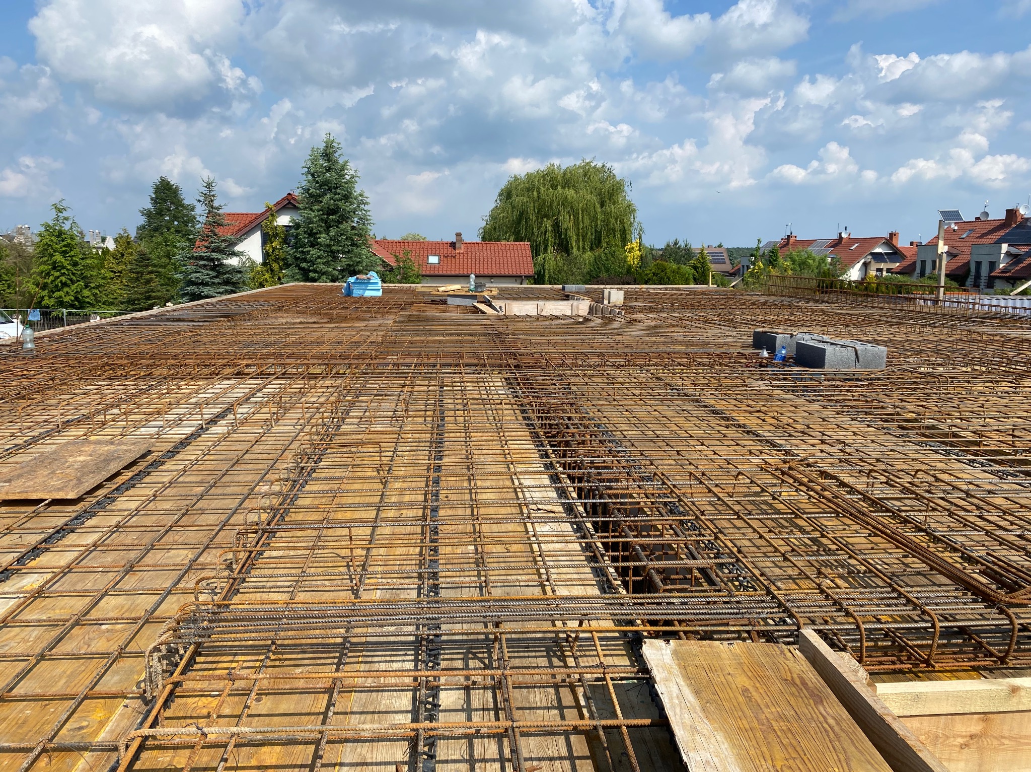 Zbrojenie stropu na deskowaniu, widok z góry na przygotowaną konstrukcję z prętów stalowych, w tle zabudowa jednorodzinna i błękitne niebo z chmurami.