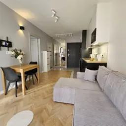 Jasne wnętrze mieszkania z drewnianą podłogą ułożoną w jodełkę, szarym narożnikiem z poduszką, drewnianym stołem z kwiatami i czarnymi krzesłami, oraz aneksem kuchennym z czarnymi szafkami.