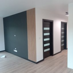 Sur-Bud - Nowoczesne wnętrze z ciemną ścianą, drewnianymi lamelami, dwiema ciemnymi drzwiami z przeszkleniami i jasną podłogą z paneli.