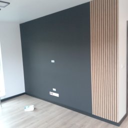 Sur-Bud - Pomalowana na ciemno szara ściana z drewnianymi lamelami po prawej stronie, wykończona czarną listwą przypodłogową, z widocznymi gniazdkami i włącznikami oraz fragmentem podłogi wyłożonej panelami.