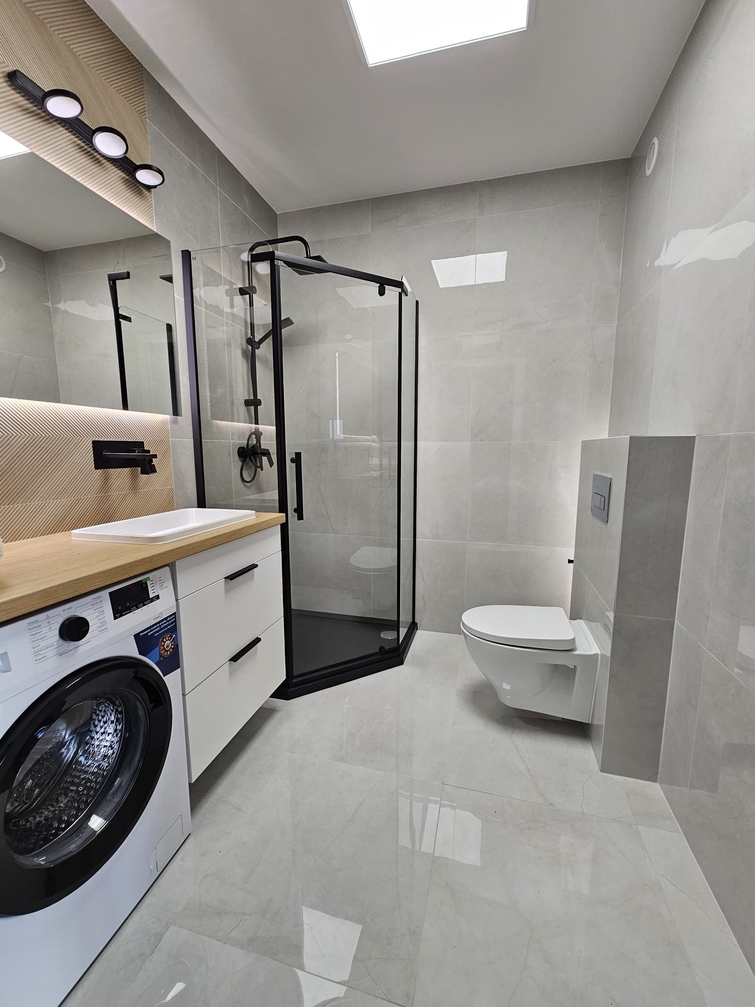 Nowoczesna łazienka z szarymi płytkami, kabiną prysznicową z czarnymi ramami, białą umywalką na drewnianym blacie, pralką i podwieszaną toaletą.