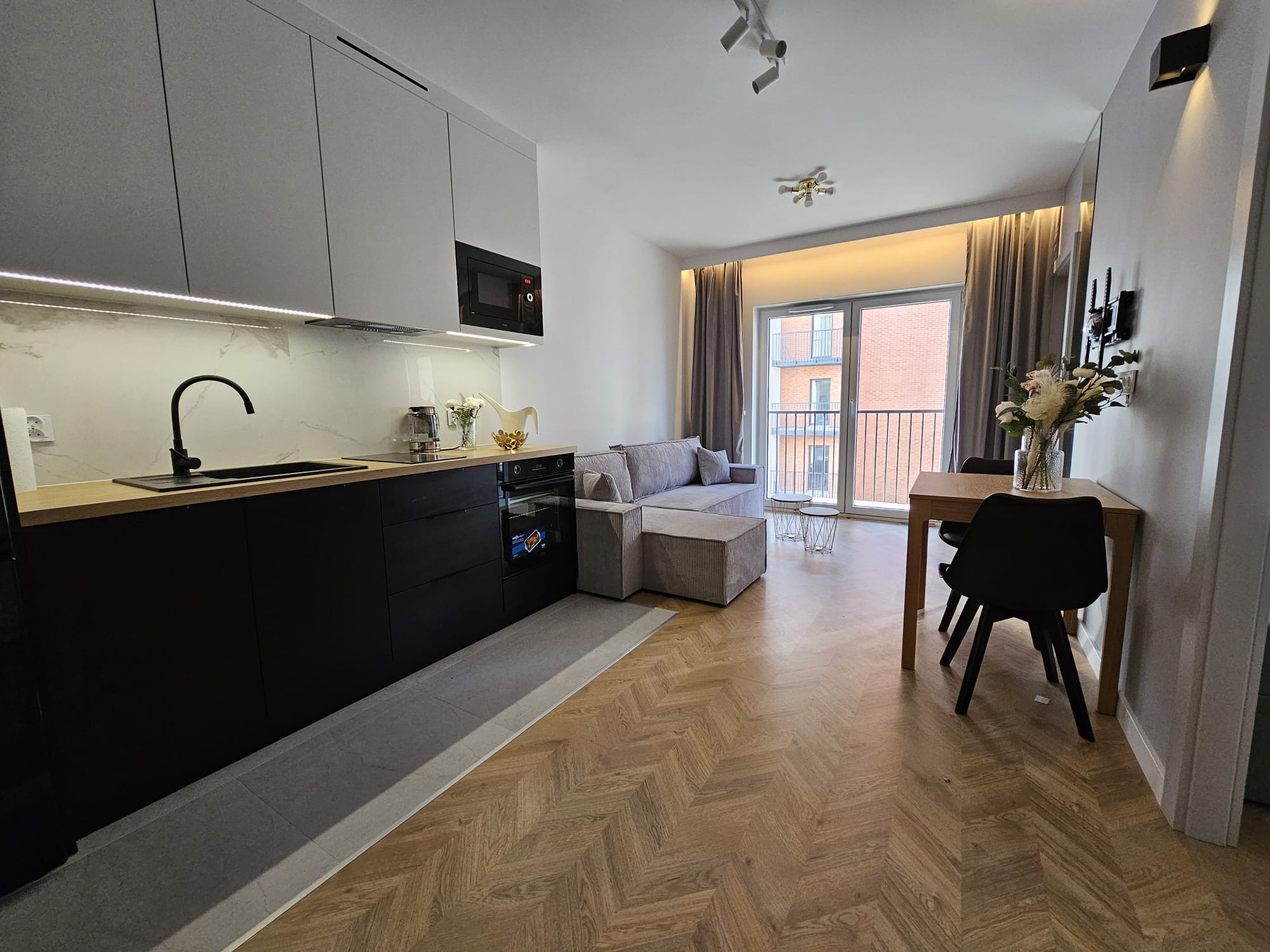 Nowoczesne wnętrze mieszkania z aneksem kuchennym, drewnianą podłogą w jodełkę, szarą sofą i balkonem z widokiem na ceglany budynek.