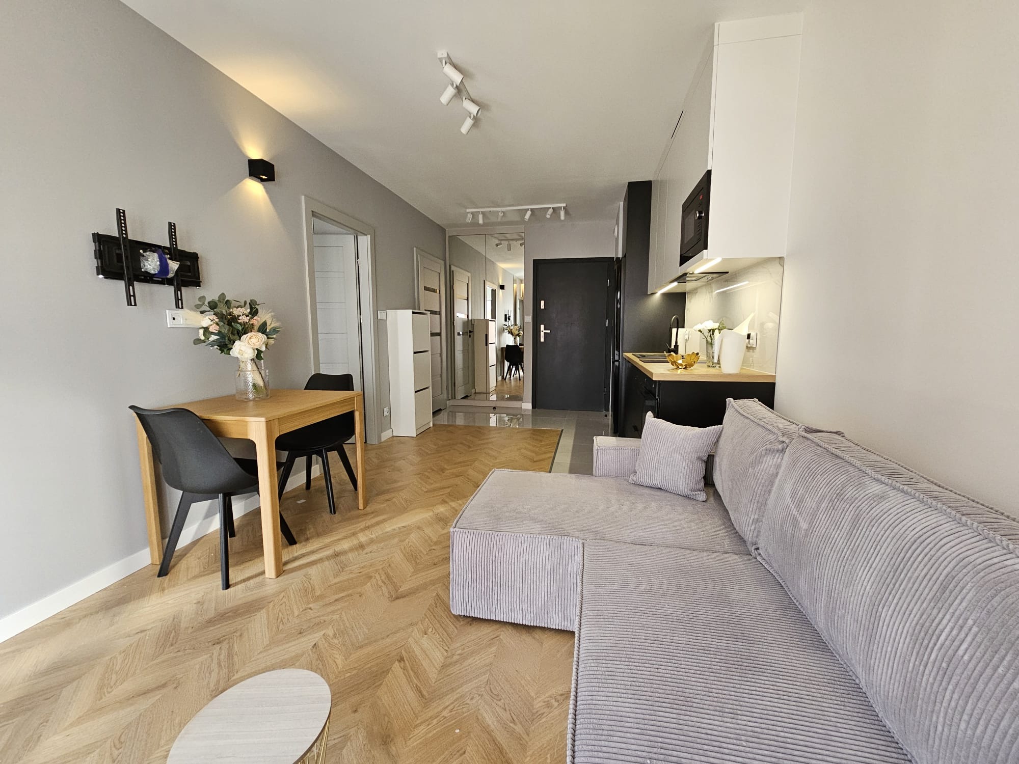 Jasne wnętrze mieszkania z drewnianą podłogą ułożoną w jodełkę, szarym narożnikiem z poduszką, drewnianym stołem z kwiatami i czarnymi krzesłami, oraz aneksem kuchennym z czarnymi szafkami.