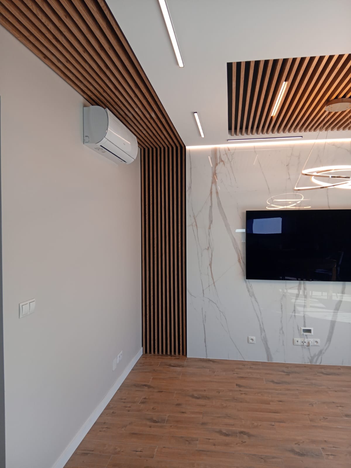 Nowoczesne wnętrze z drewnianą zabudową sufitu i ściany, marmurową ścianą z telewizorem oraz parkietem, oświetlone liniowymi lampami LED.