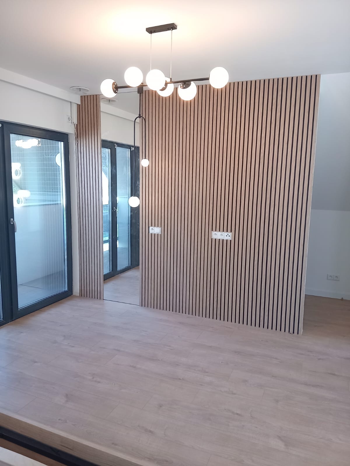 Nowoczesne wnętrze z drewnianymi lamelami na ścianie, jasną podłogą panelową i designerską lampą sufitową z kulistymi kloszami, widok na okno balkonowe.