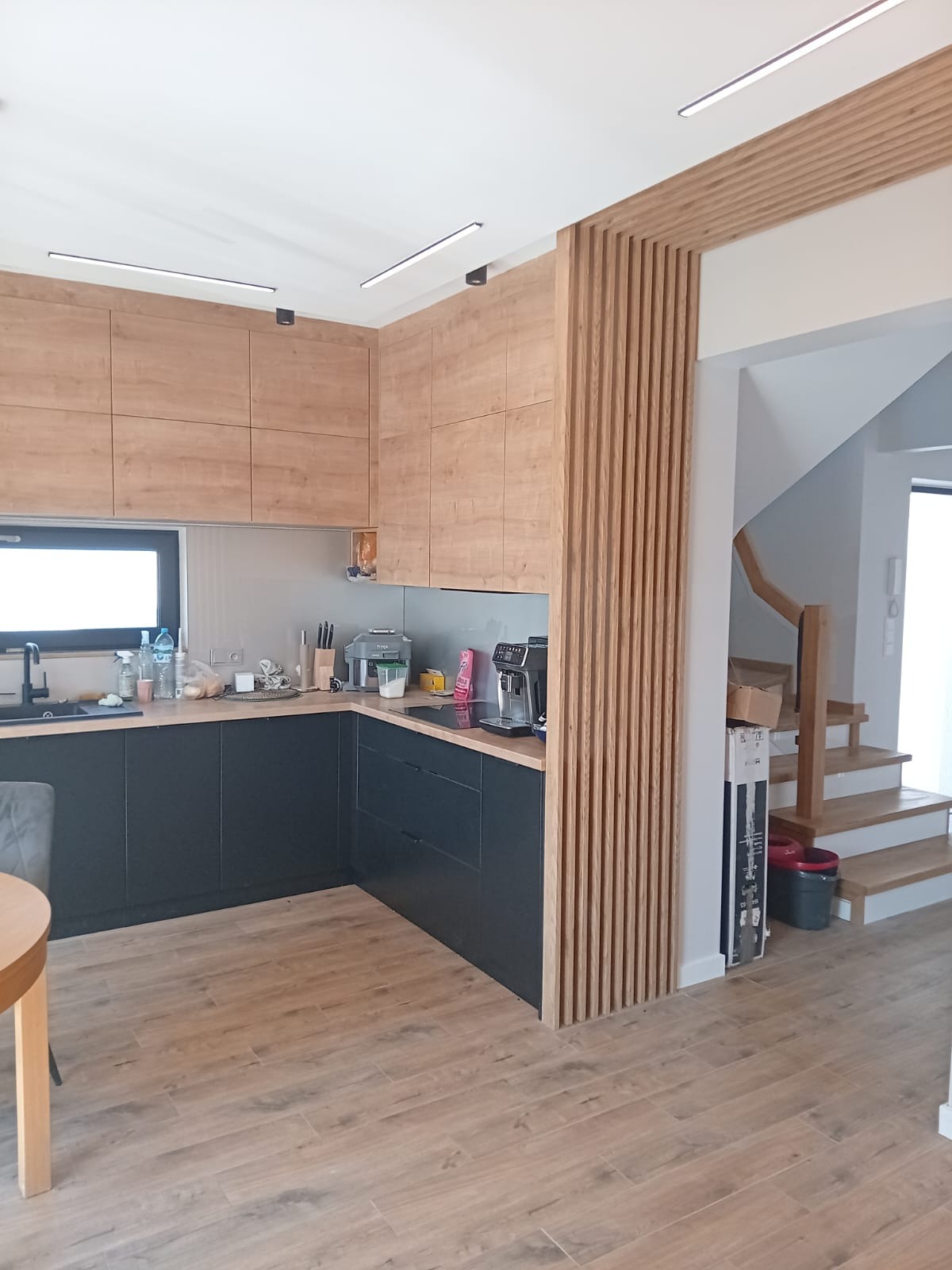 Nowoczesna kuchnia z drewnianymi szafkami górnymi, czarnymi szafkami dolnymi i drewnianą ścianką działową z pionowych listew, widok na drewniane schody w tle.