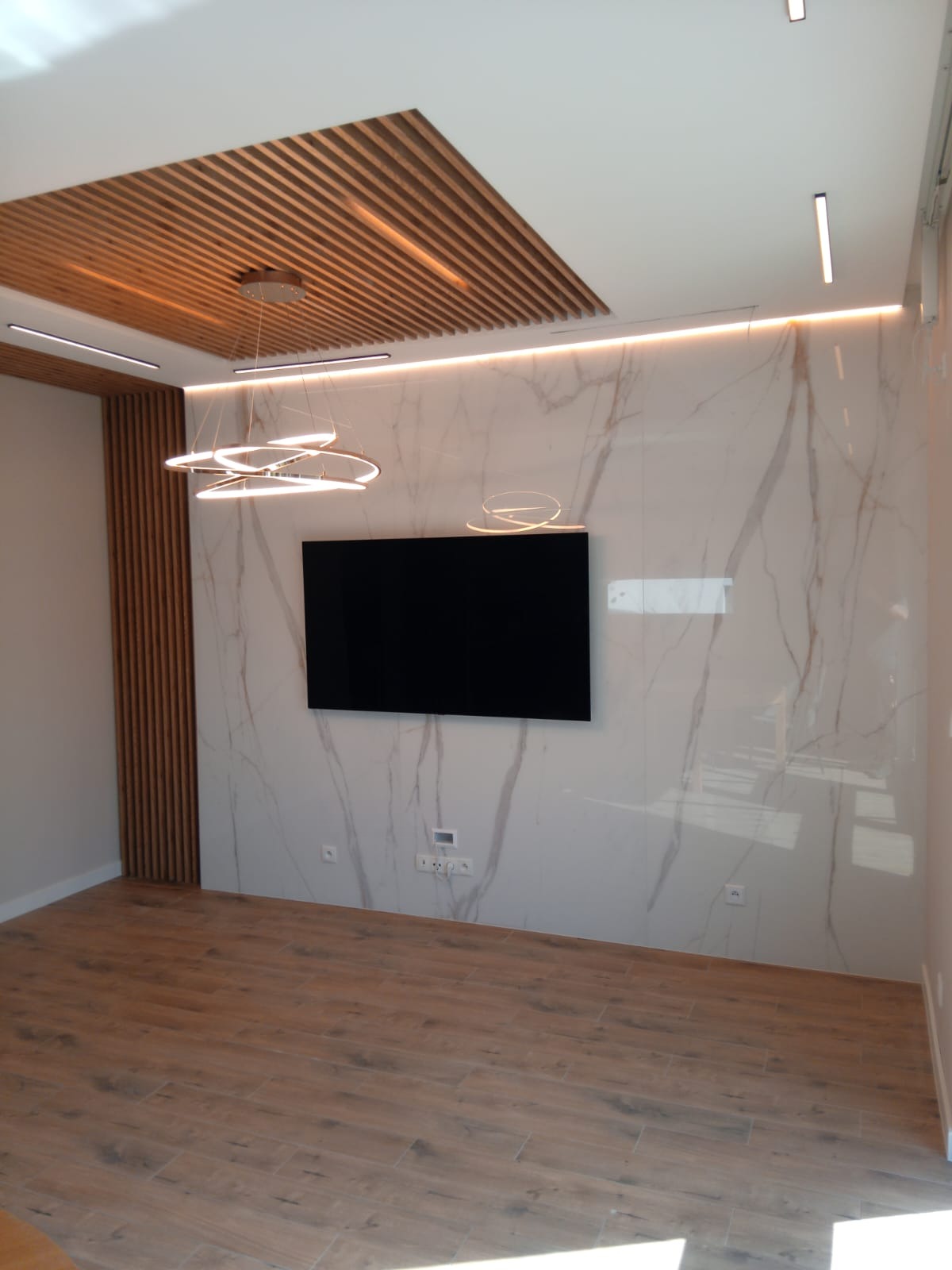 Nowoczesny salon z drewnianą zabudową sufitu i ściany, podświetlaną listwą LED, marmurową ścianą z telewizorem i drewnopodobną podłogą z gresu.