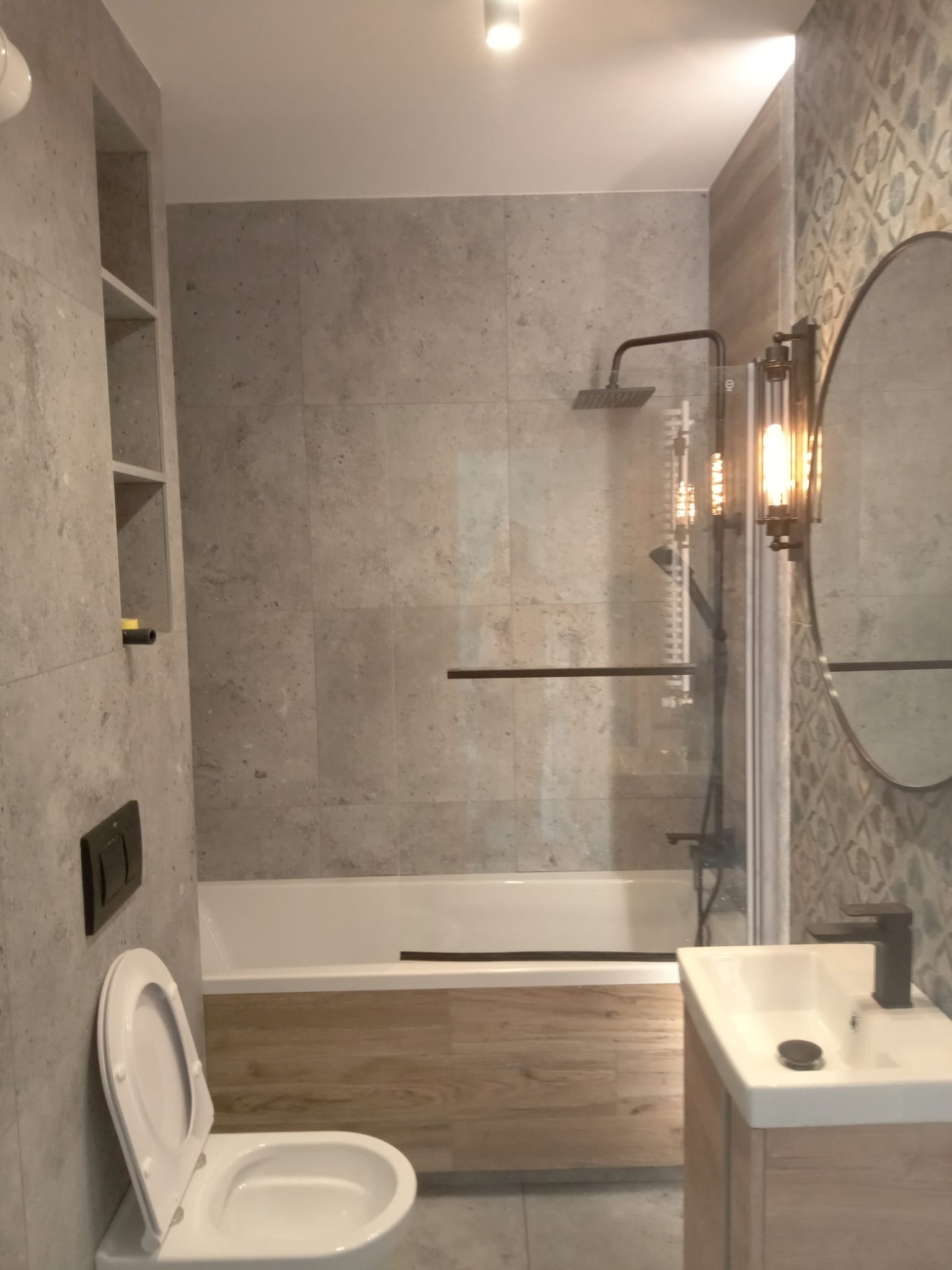 Nowoczesna łazienka z szarą płytką imitującą kamień, kabiną prysznicową typu walk-in z czarną armaturą, podłogą z paneli drewnopodobnych, owalnym lustrem z dekoracyjną lampą i toaletą z otwartą klapą.