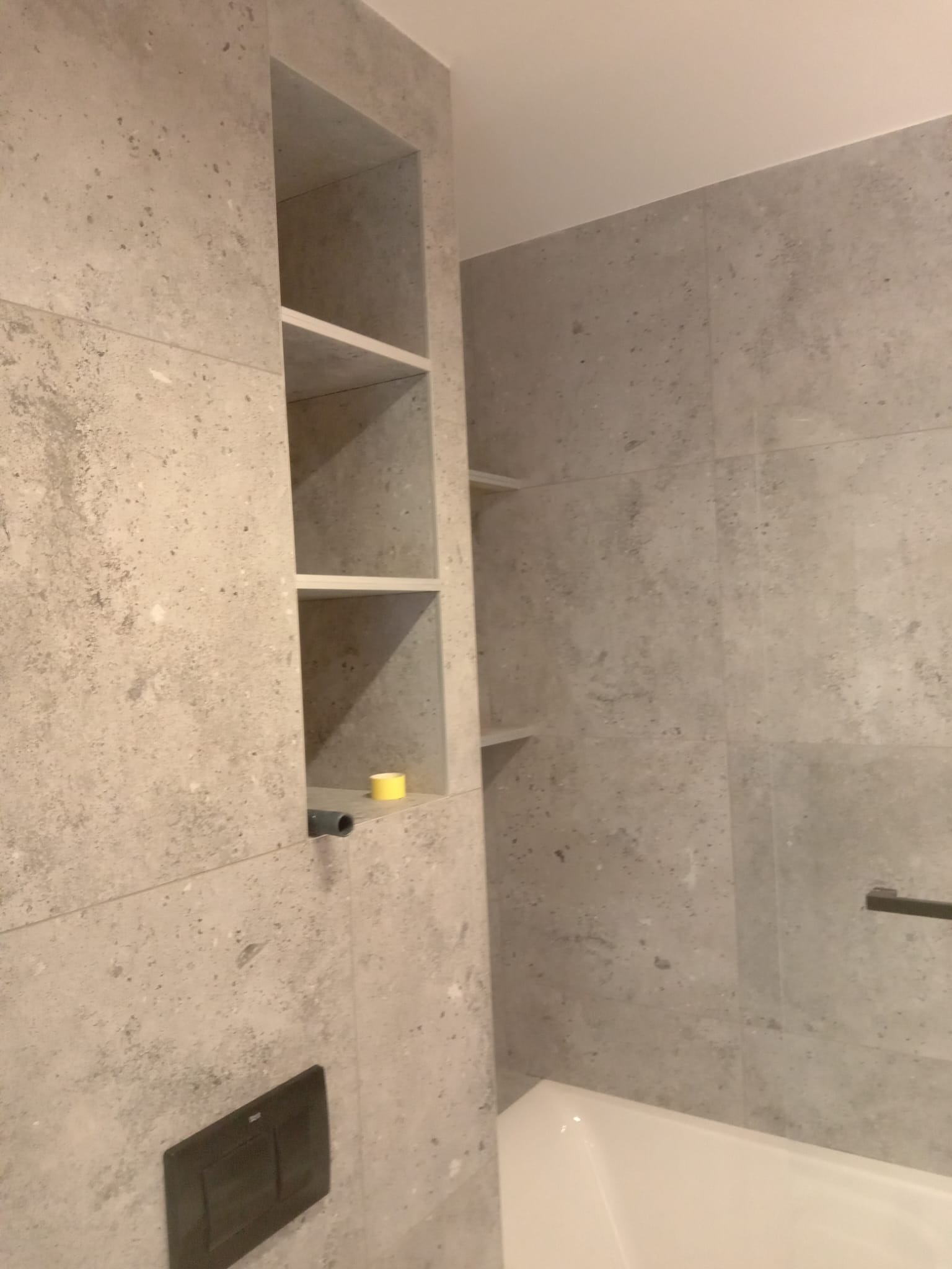 Ściana łazienki wyłożona szarymi płytkami imitującymi kamień, z wnęką z półkami, widoczny fragment wanny i czarny przycisk spłuczki.