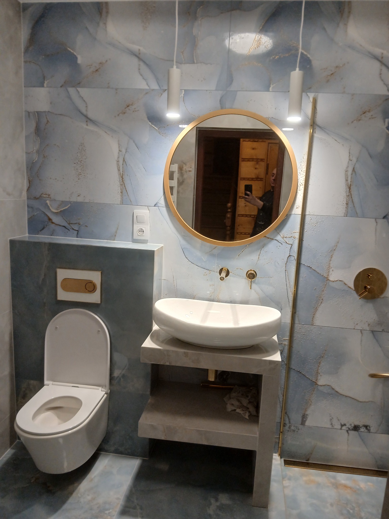 Nowoczesna łazienka z marmurowymi płytkami w odcieniach błękitu i szarości, okrągłe lustro w złotej ramie, biała umywalka nablatowa, wisząca toaleta i minimalistyczne oświetlenie.